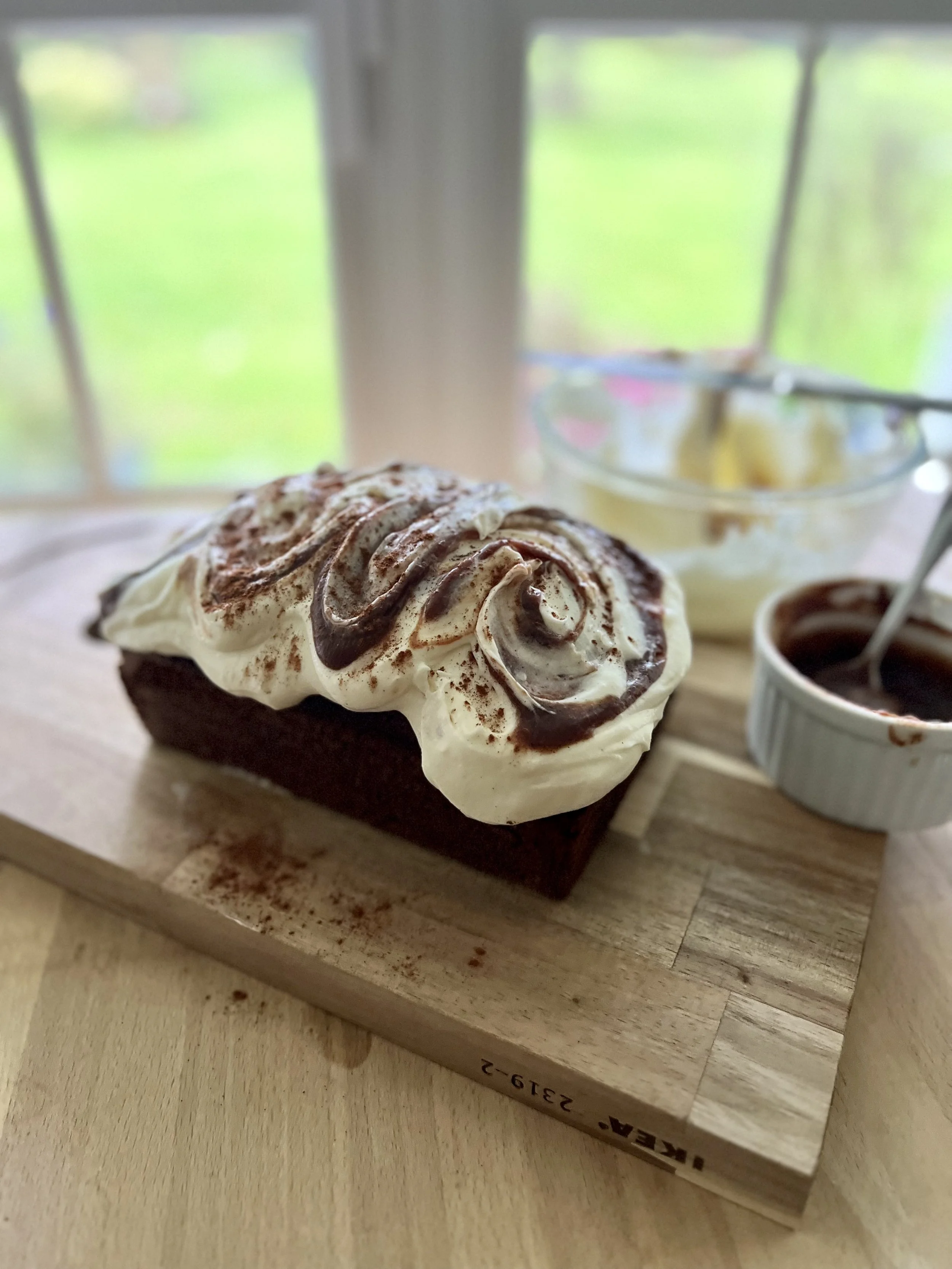 A wooden cutting board holding a large piece of whipped cream-topped dessert with chocolate drizzle, next to small bowls of chocolate sauce and whipped cream, in front of a blurred window with greenery outside.