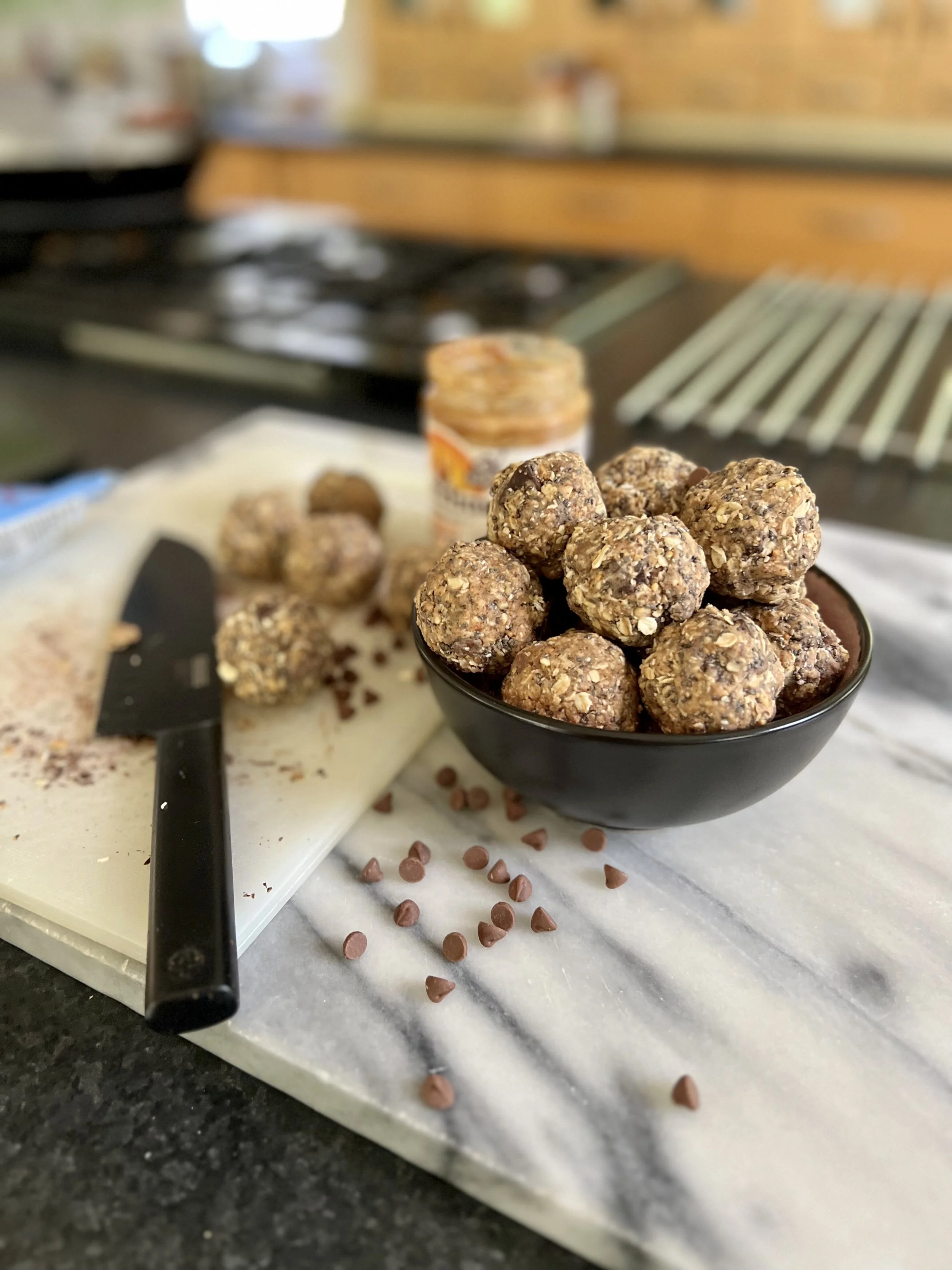 Close-up of homemade energy bites on a black bowl, with chocolate chips scattered on a white surface, a butter knife, and a jar of nut spread in the kitchen.