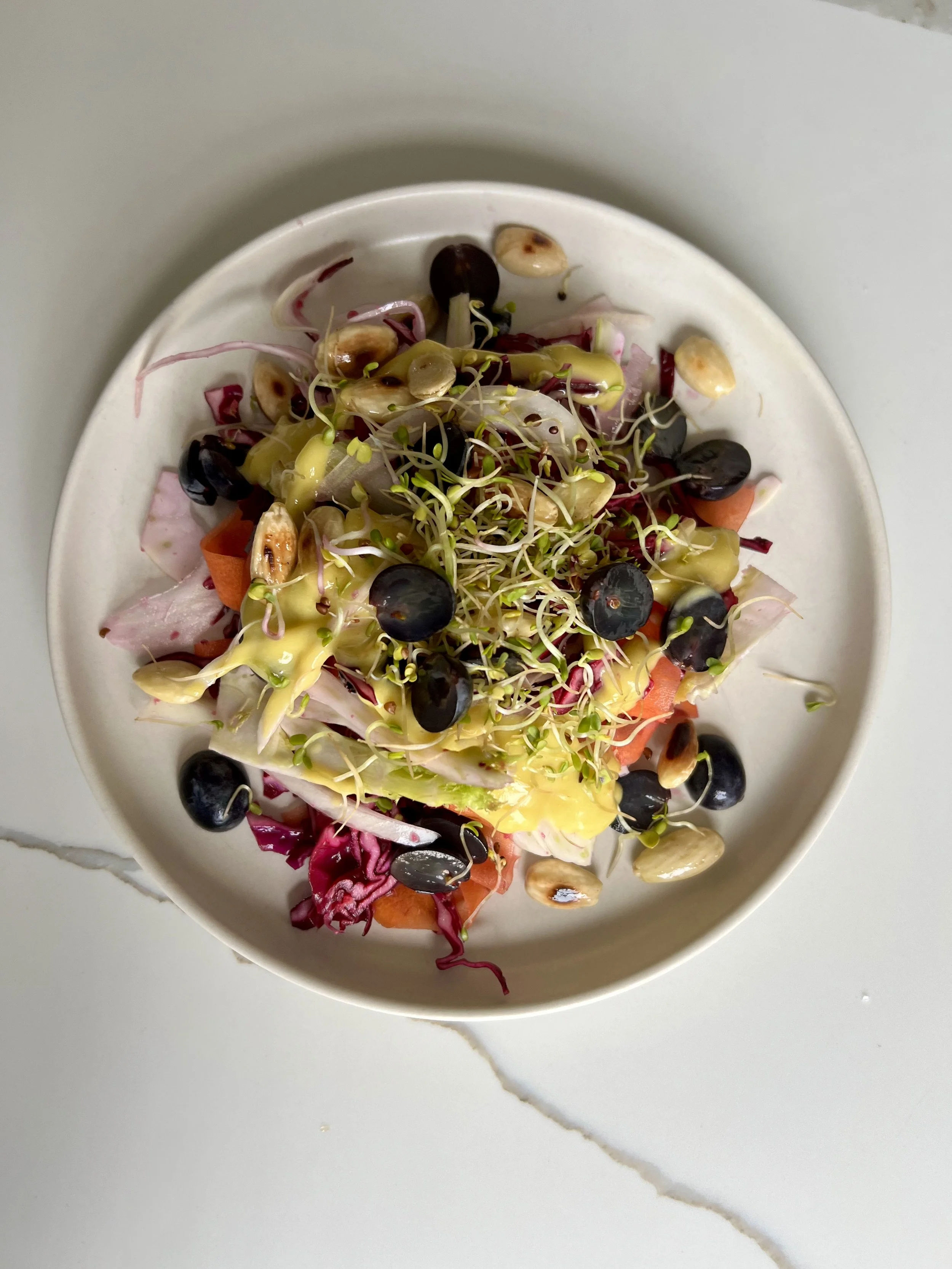 Assorted salad with sprouts, blueberries, shredded vegetables, and mixed greens in a white bowl on a light gray marble surface.