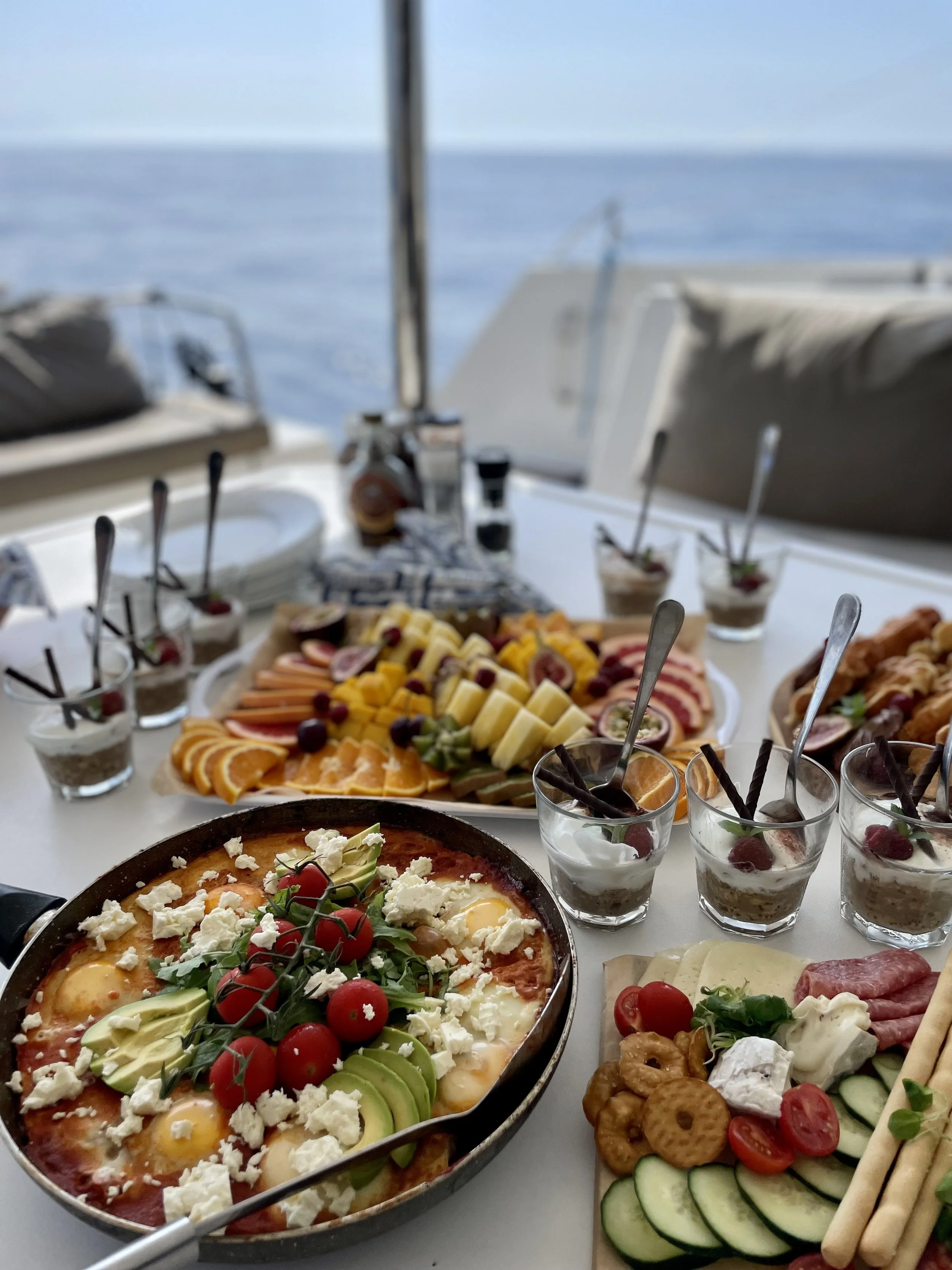 A table with a variety of foods including a frittata with eggs, tomatoes, and cheese, a vegetable platter with cucumber, cherry tomatoes, and crackers, bowls of parfait with whipped cream and berries, and a large platter of assorted fresh fruit inclu