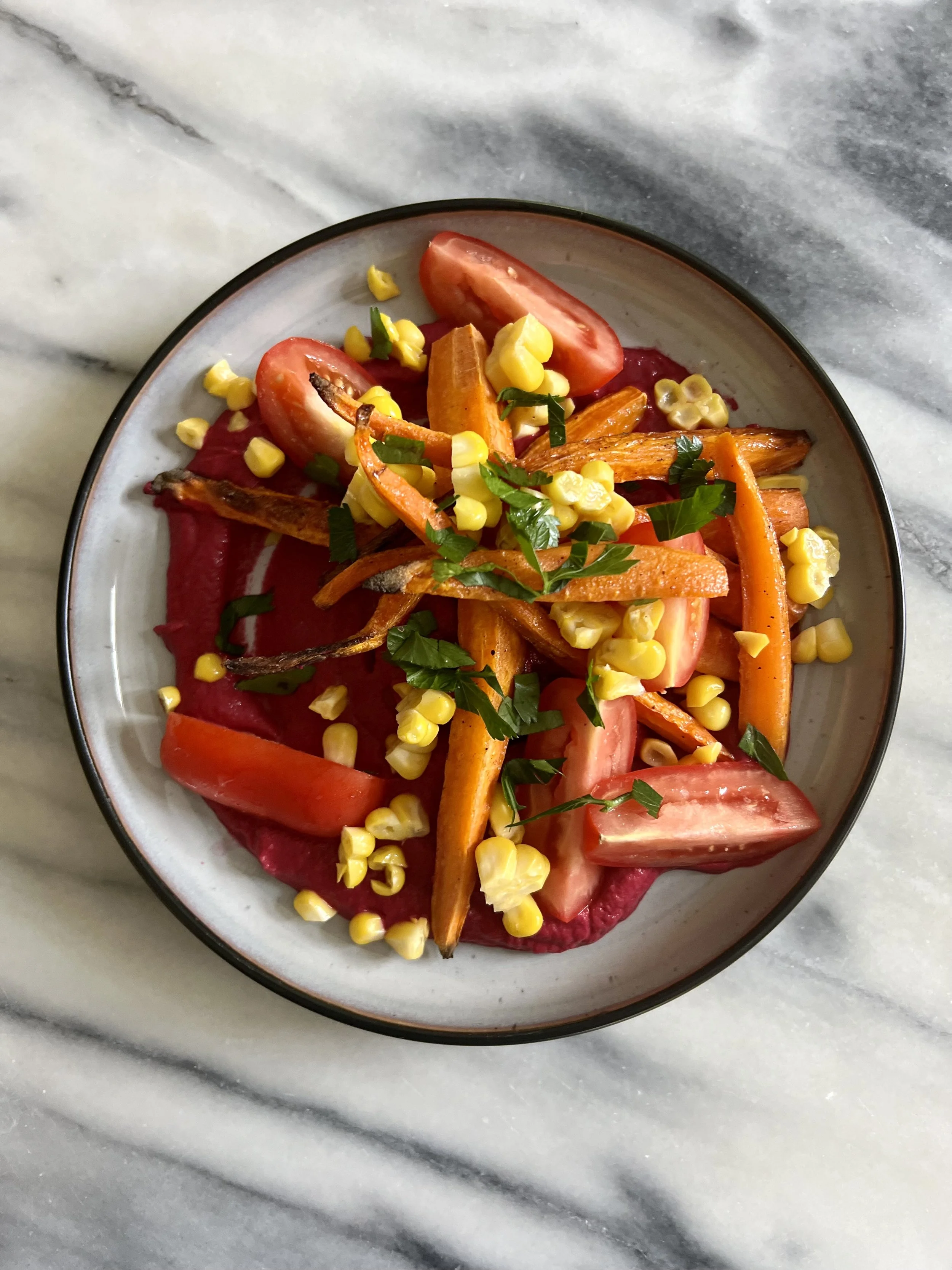 A plate of roasted vegetables including cherry tomatoes, carrots, corn, and herbs, set on a marble surface.