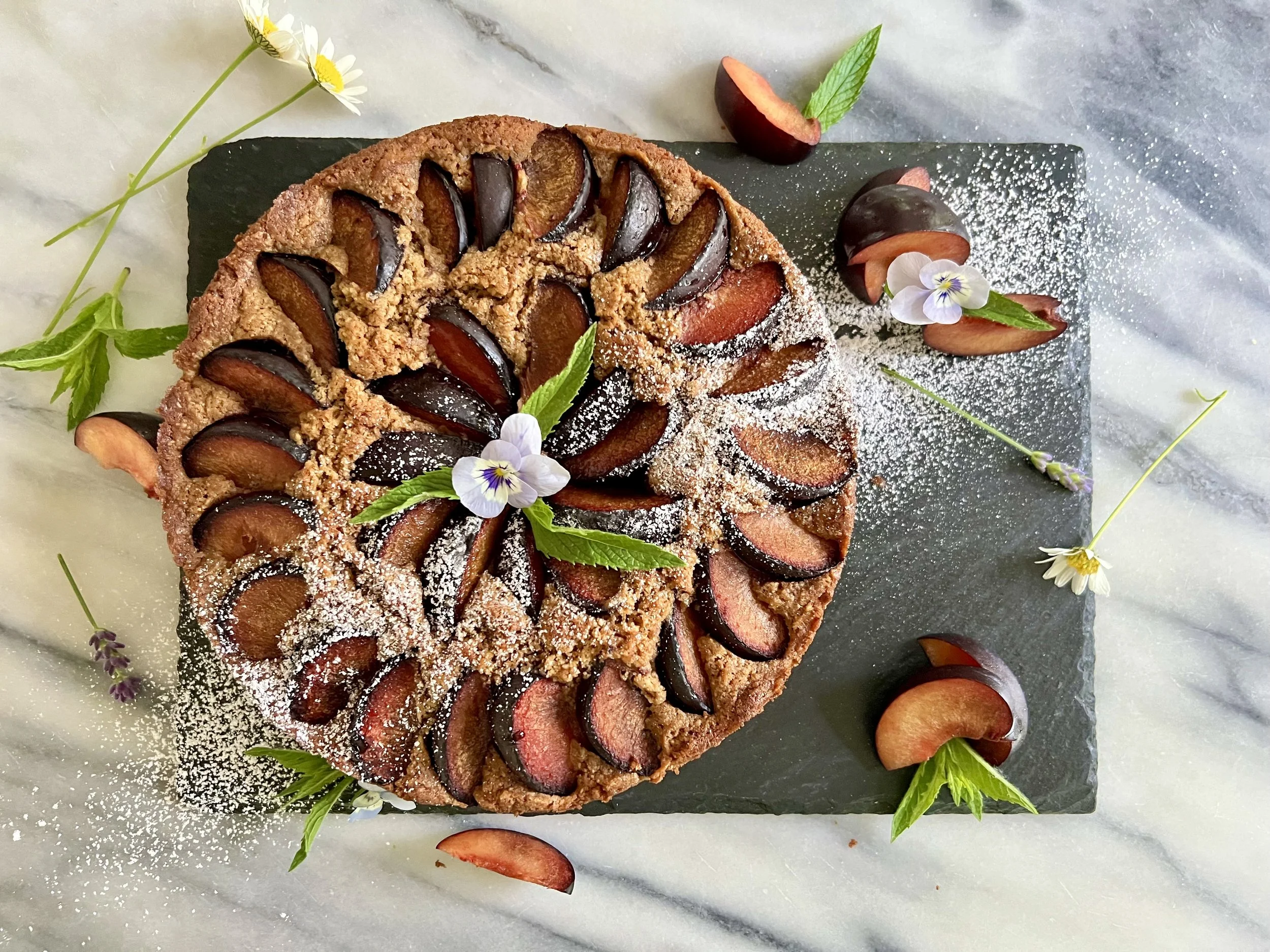 Sliced plum tart garnished with mint leaves and edible flowers, dusted with powdered sugar, on a marble surface.