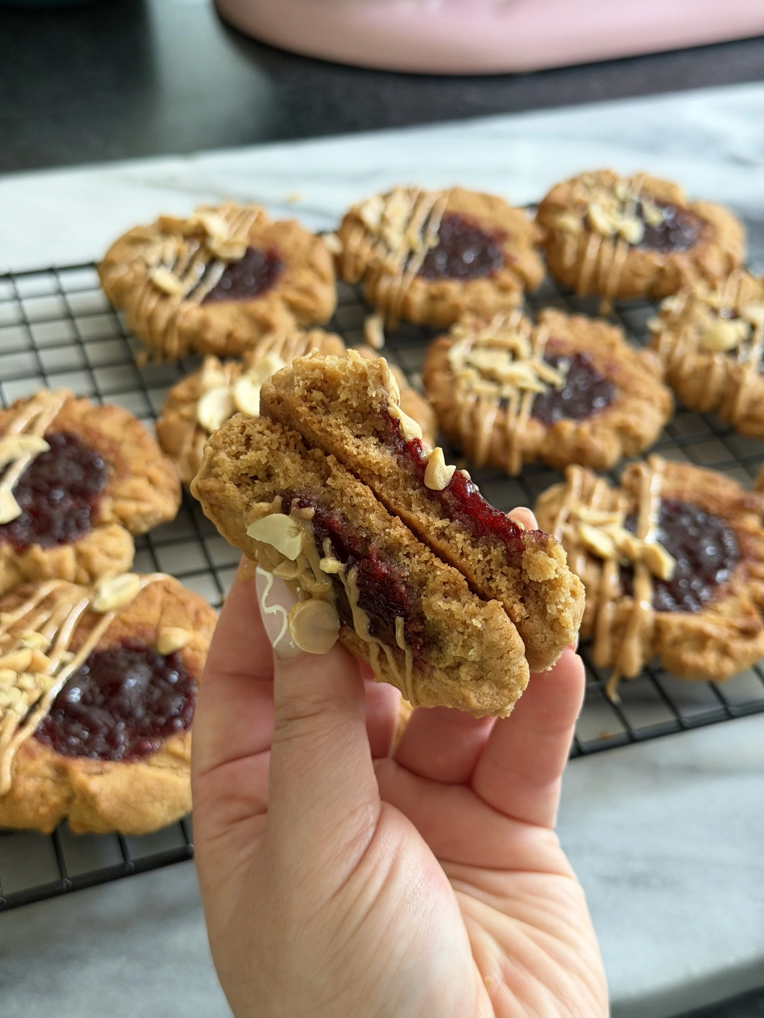 Close-up of a hand holding a bitten oatmeal cookie with cherry filling and white chocolate chips. In the background, there is a wire cooling rack with several similar cookies topped with drizzled white chocolate and chopped nuts, on a gray surface.