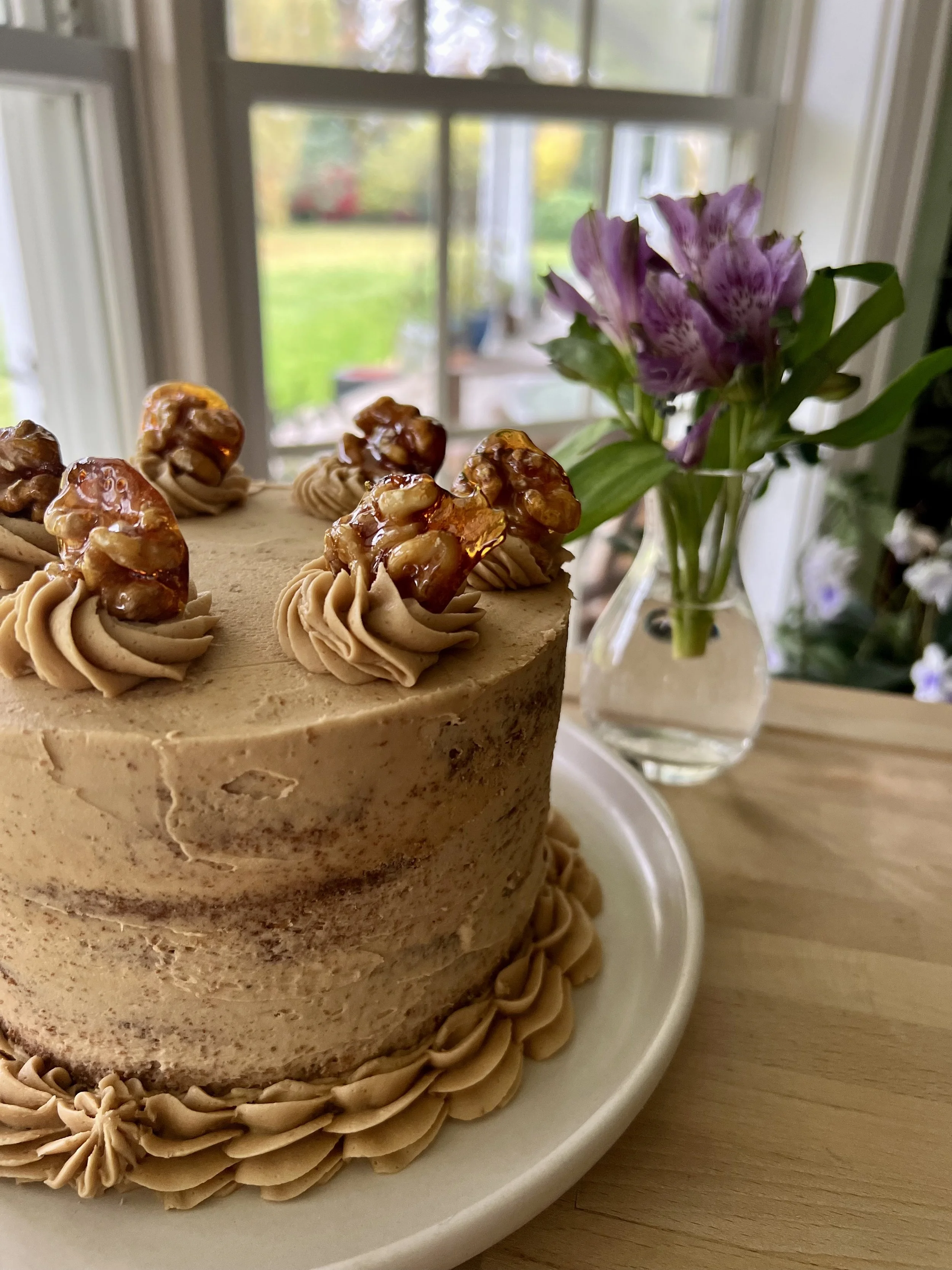 A close-up image of a beige or light brown cake decorated with caramelized nuts on top and piped frosting around the base, placed on a white plate on a wooden surface. In the background, there is a vase with purple and white flowers and a window with