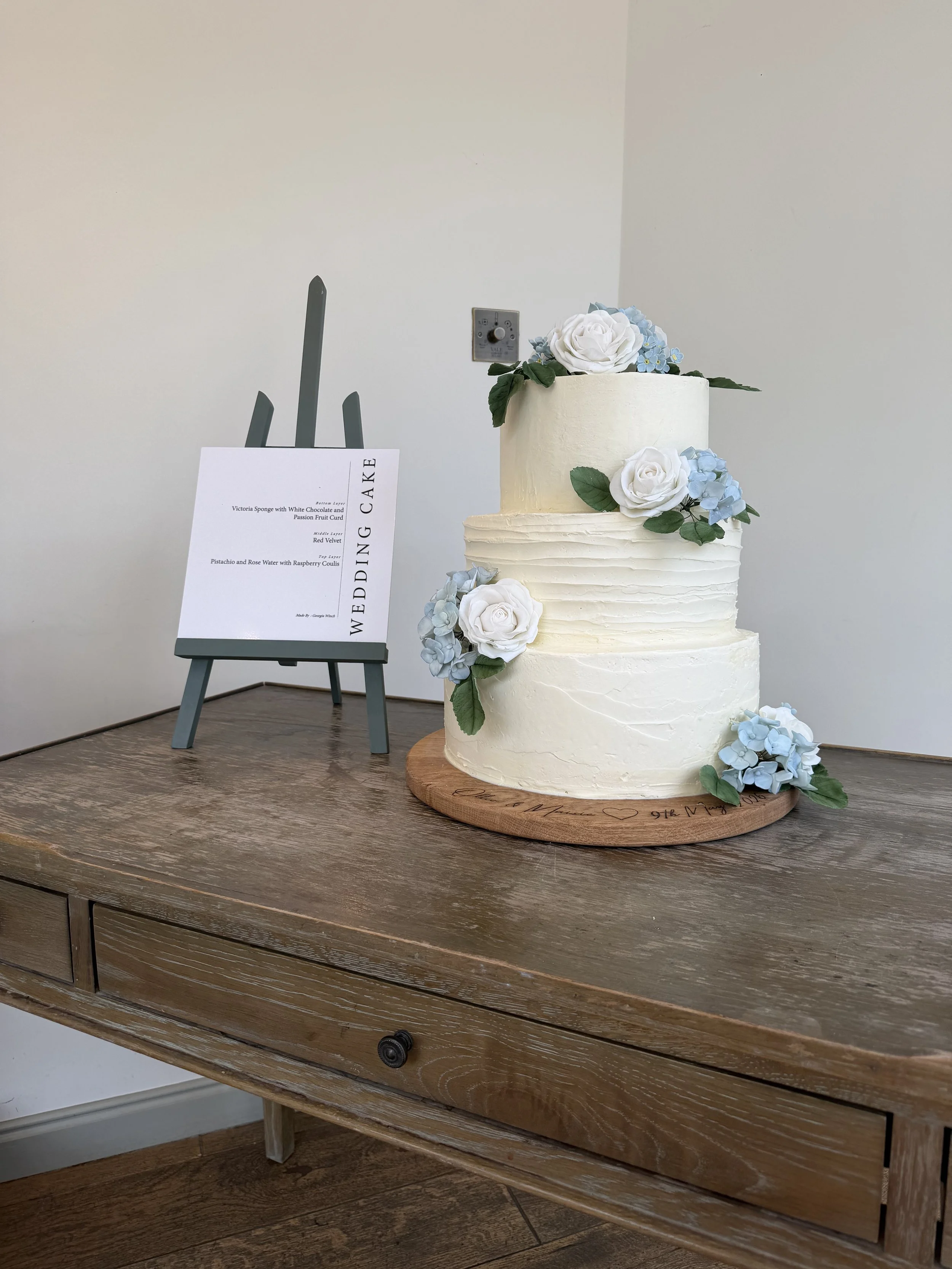 A three-tier white wedding cake decorated with white roses, light blue hydrangeas, and green leaves, placed on a wooden table with a handwritten message on the base.