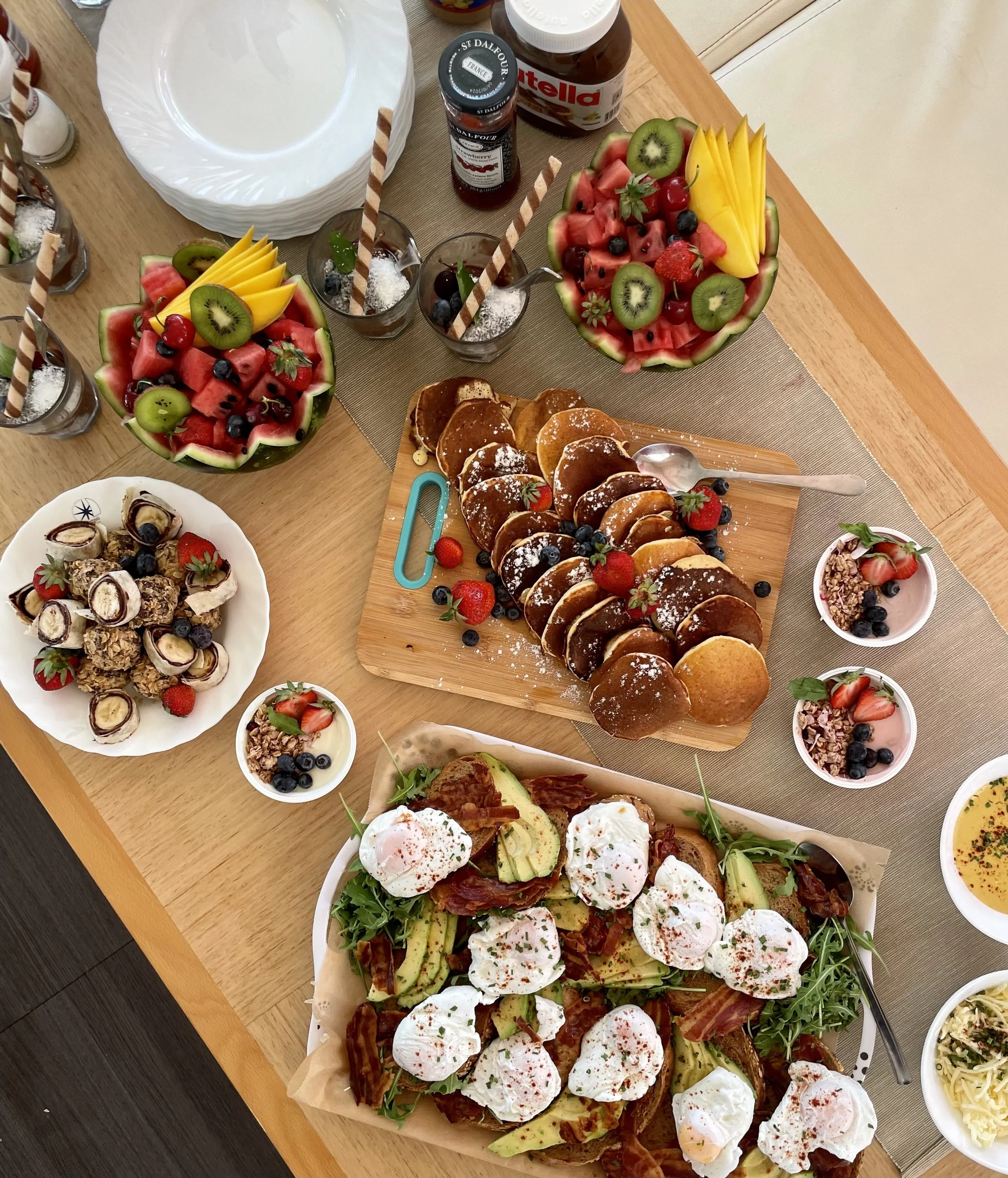 Assorted dishes on a wooden table including bowls of yogurt with poached eggs, a large platter of open-faced sandwiches topped with poached eggs, bacon, avocado, and ham, a sliced pancake or waffle with berries, a large watermelon bowl filled with wa
