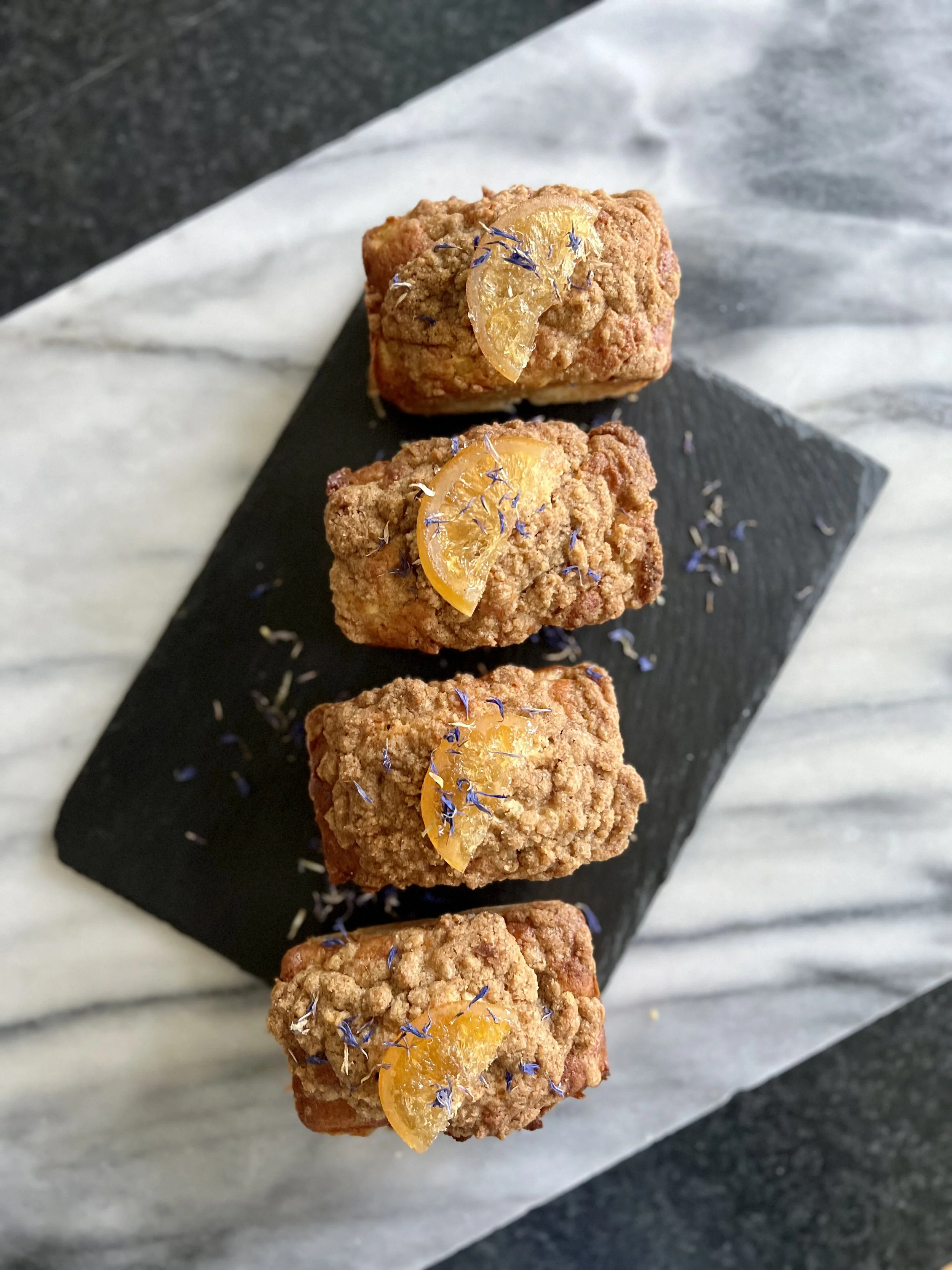 Four pieces of fried chicken topped with sliced lemon and small purple edible flowers on black slate serving platter lined with white parchment paper.