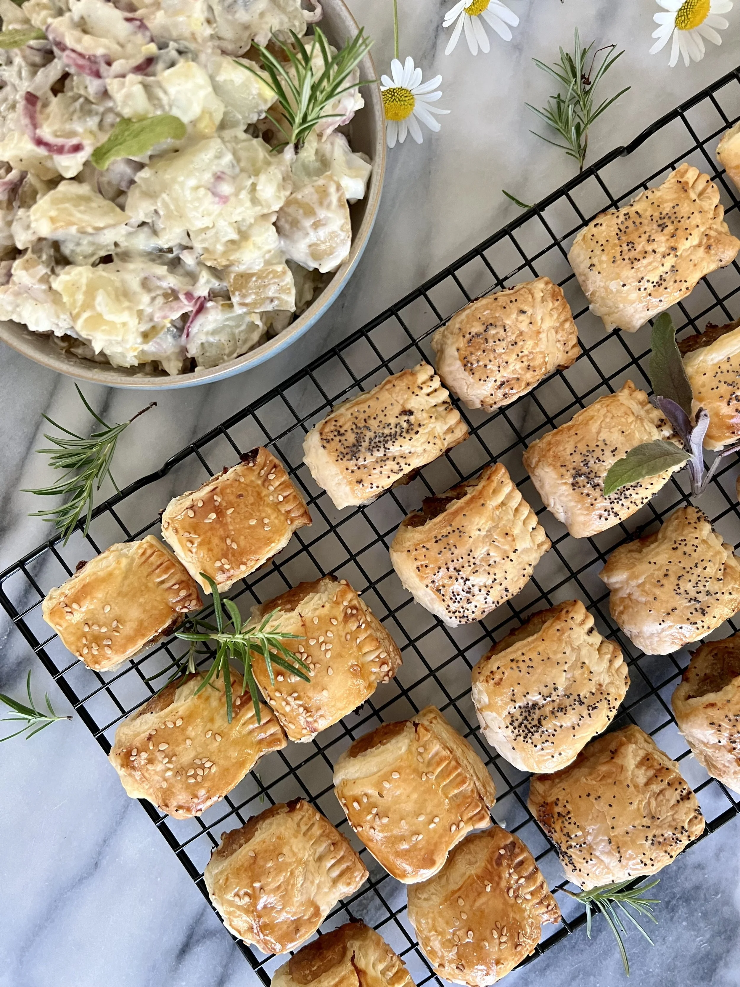 A wire cooling rack with golden, flaky puff pastry pastries topped with sesame and poppy seeds, garnished with sprigs of fresh rosemary and sage. To the upper left, a bowl of creamy potato salad with herbs, garnished with rosemary and daisies.