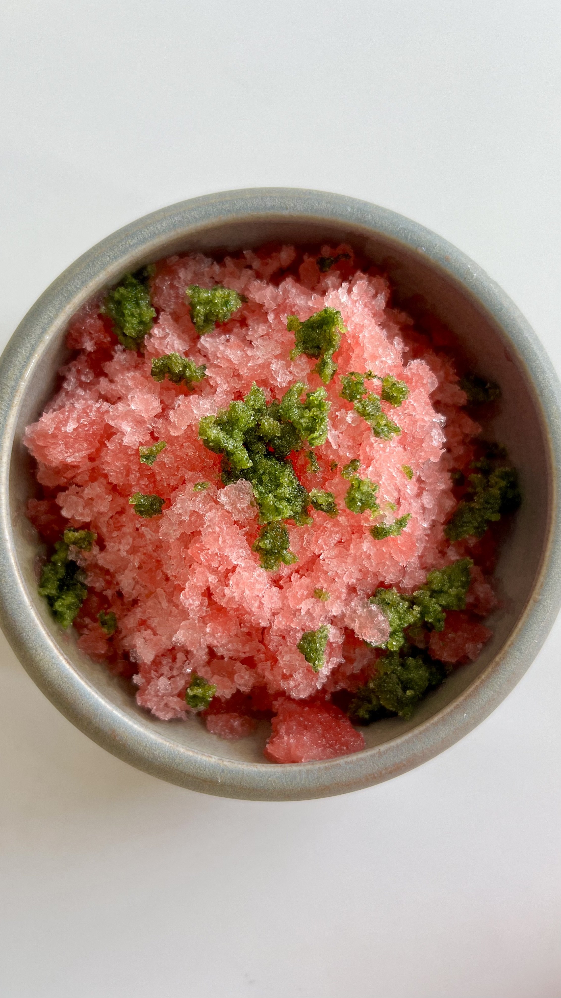 Close-up of a snow cone with pink frozen ice and green toppings in a silver cup on a white surface.