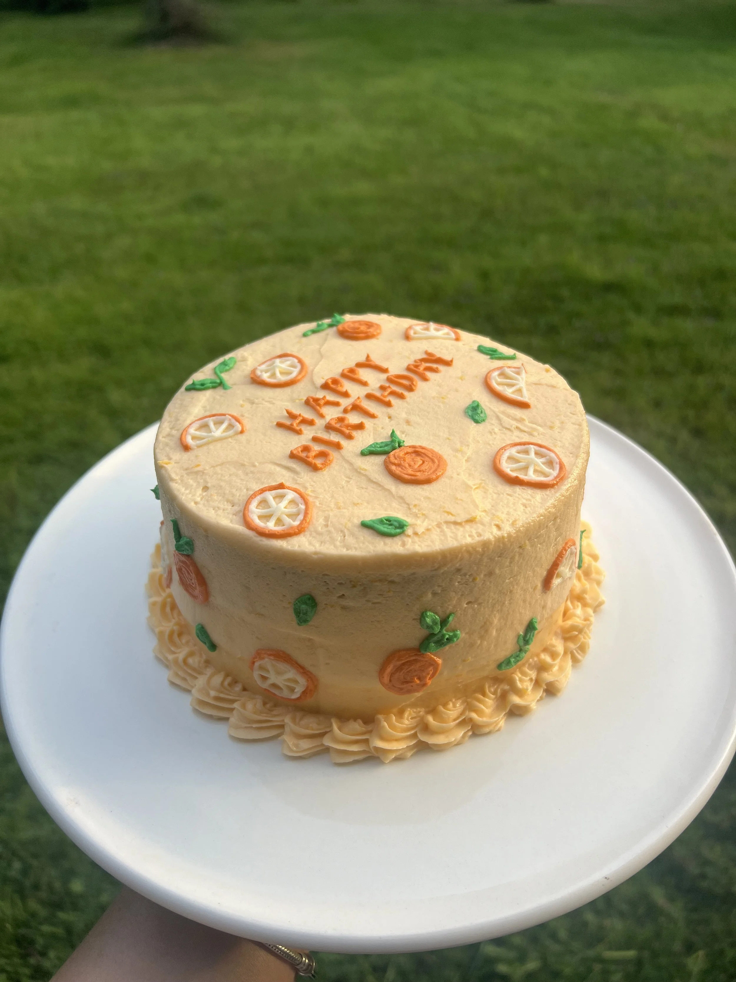 A birthday cake decorated with orange, white, and green icing, sitting on a white plate outdoors with grass in the background.