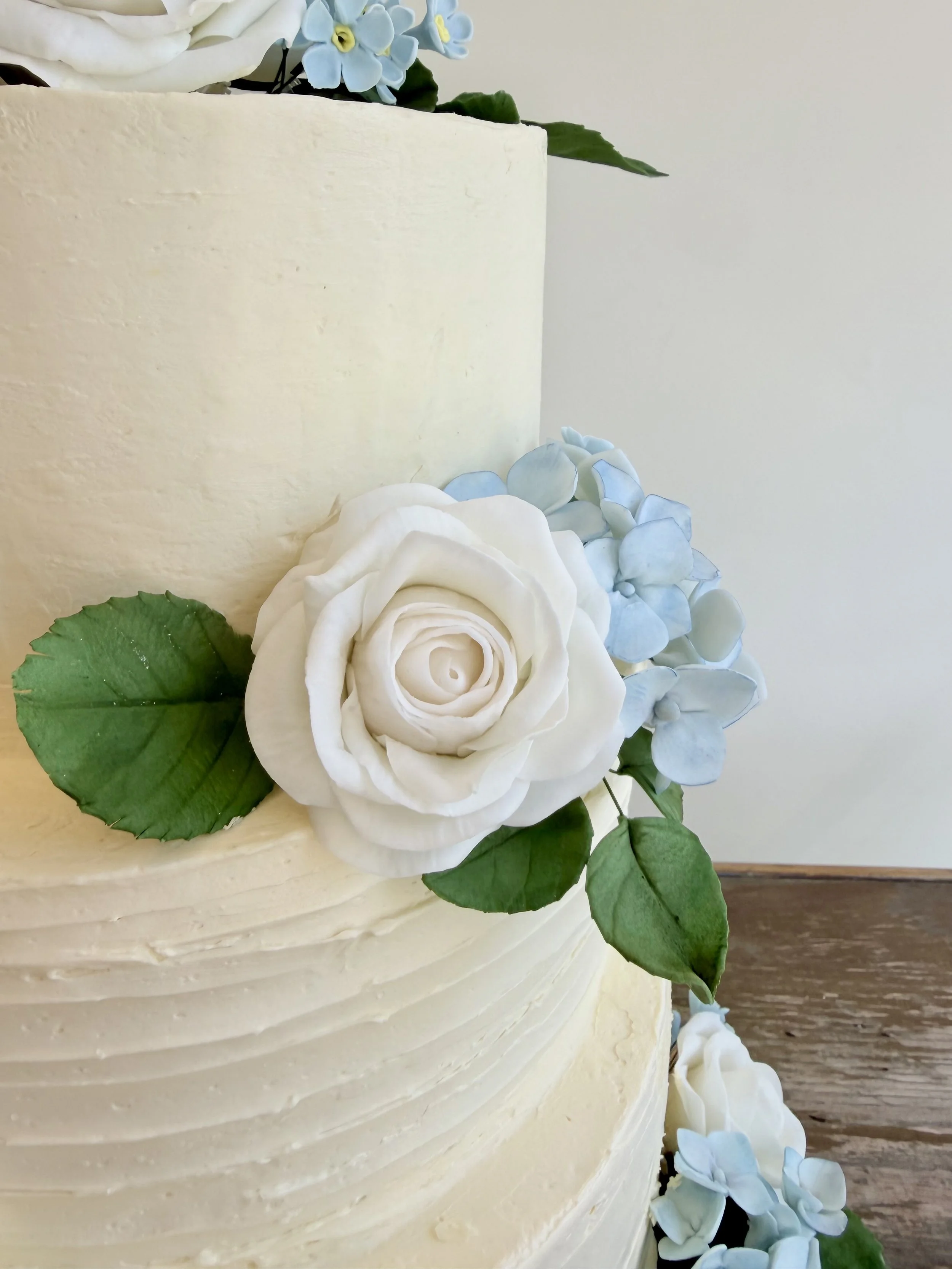 Close-up of a white wedding cake decorated with white roses, blue hydrangeas, and green leaves.