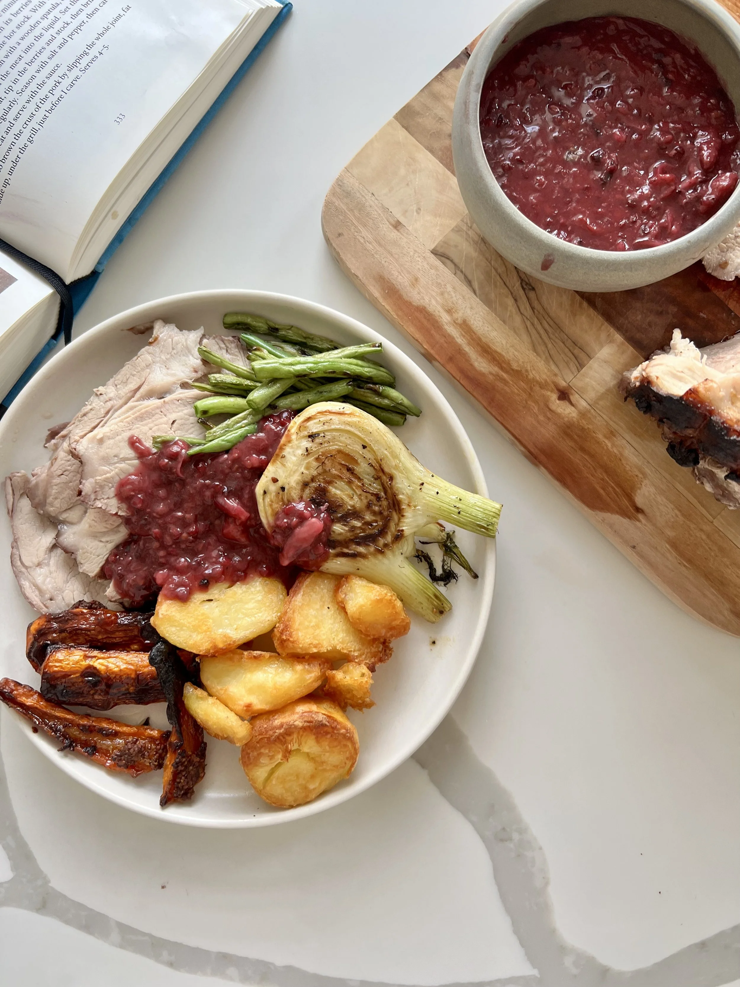 A white plate with roasted chicken, green beans, roasted lemon, and roasted potatoes on a white surface. A bowl of berry sauce and roasted meat on a wooden cutting board are also visible.