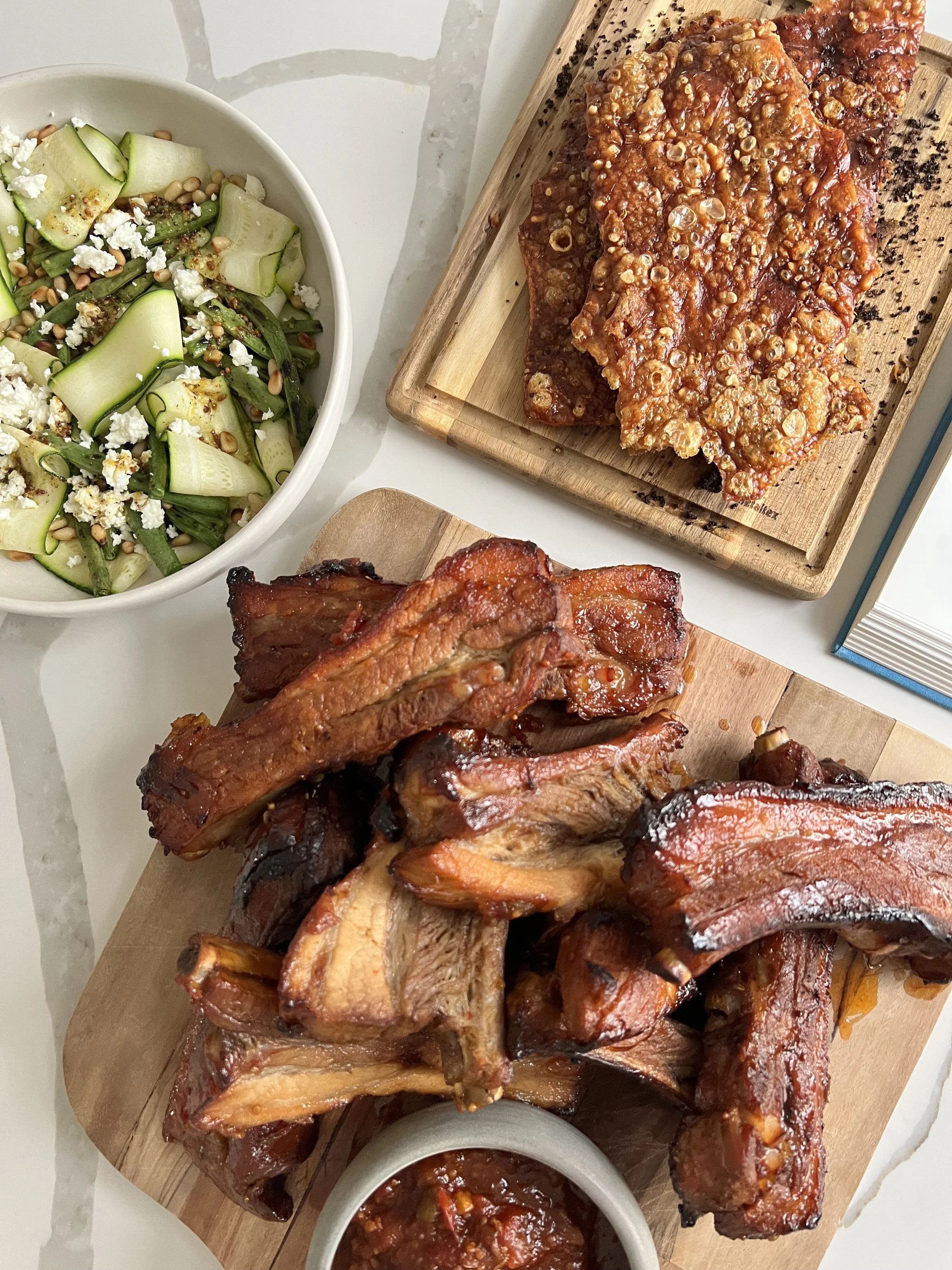 Barbecue ribs on a wooden board with sides of cucumber and feta salad, and bowls of barbecue sauce.