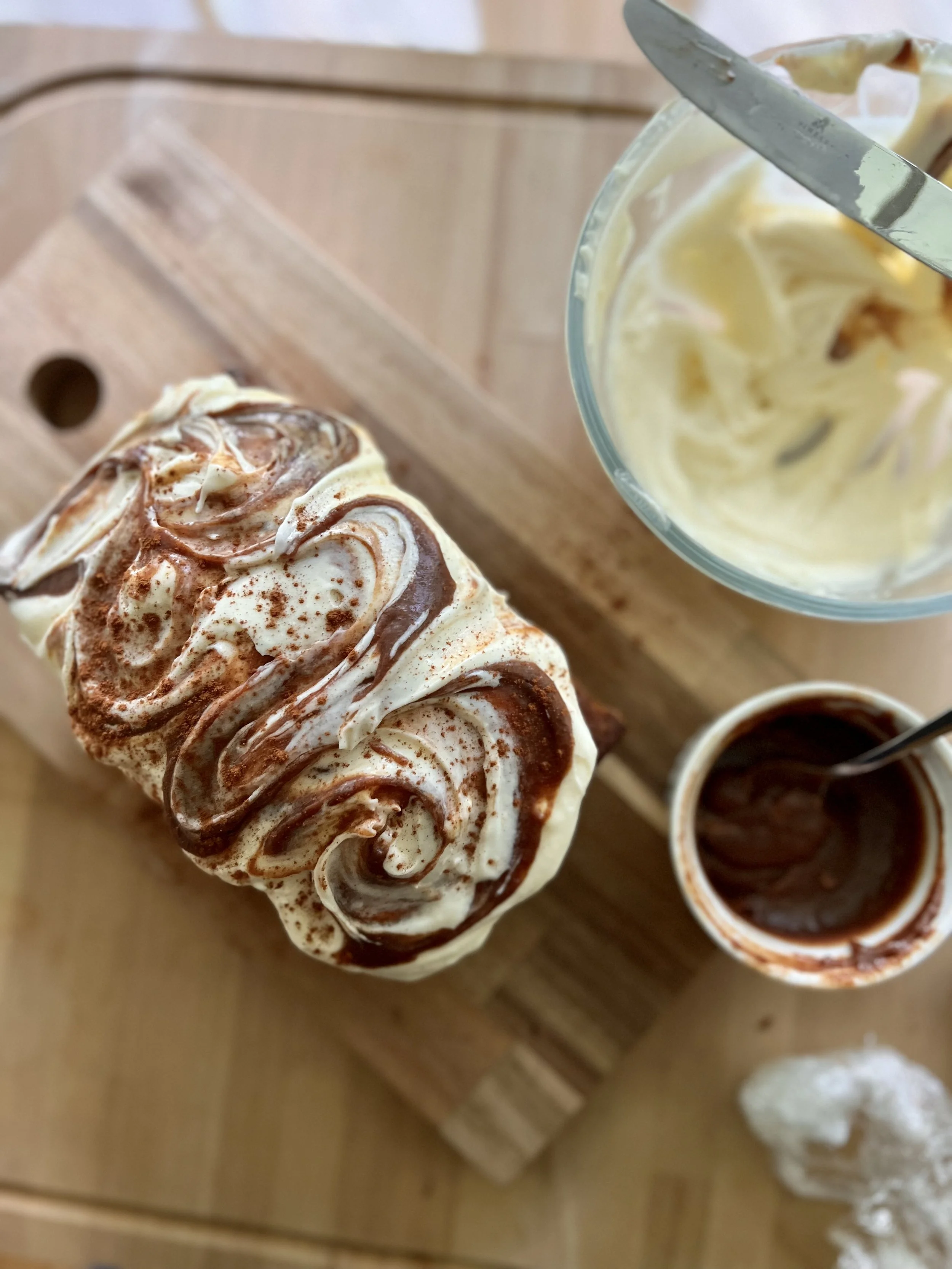 A cupcake with whipped cream and chocolate drizzle on top, placed on a wooden surface with small bowls of chocolate and additional toppings nearby.