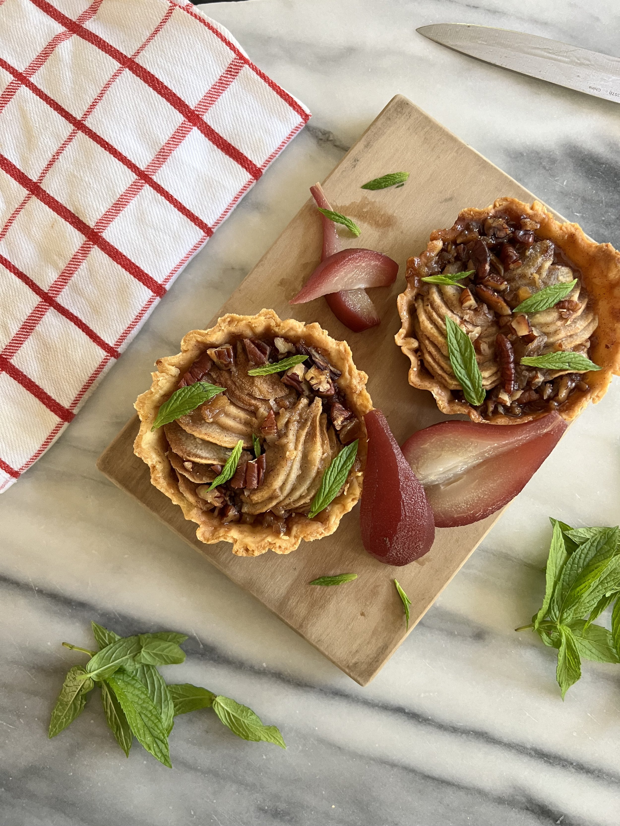 Miniature tart shells filled with pecan and caramel filling, garnished with fresh mint leaves, with slices of pear and a sprinkle of mint leaves on a marble surface.