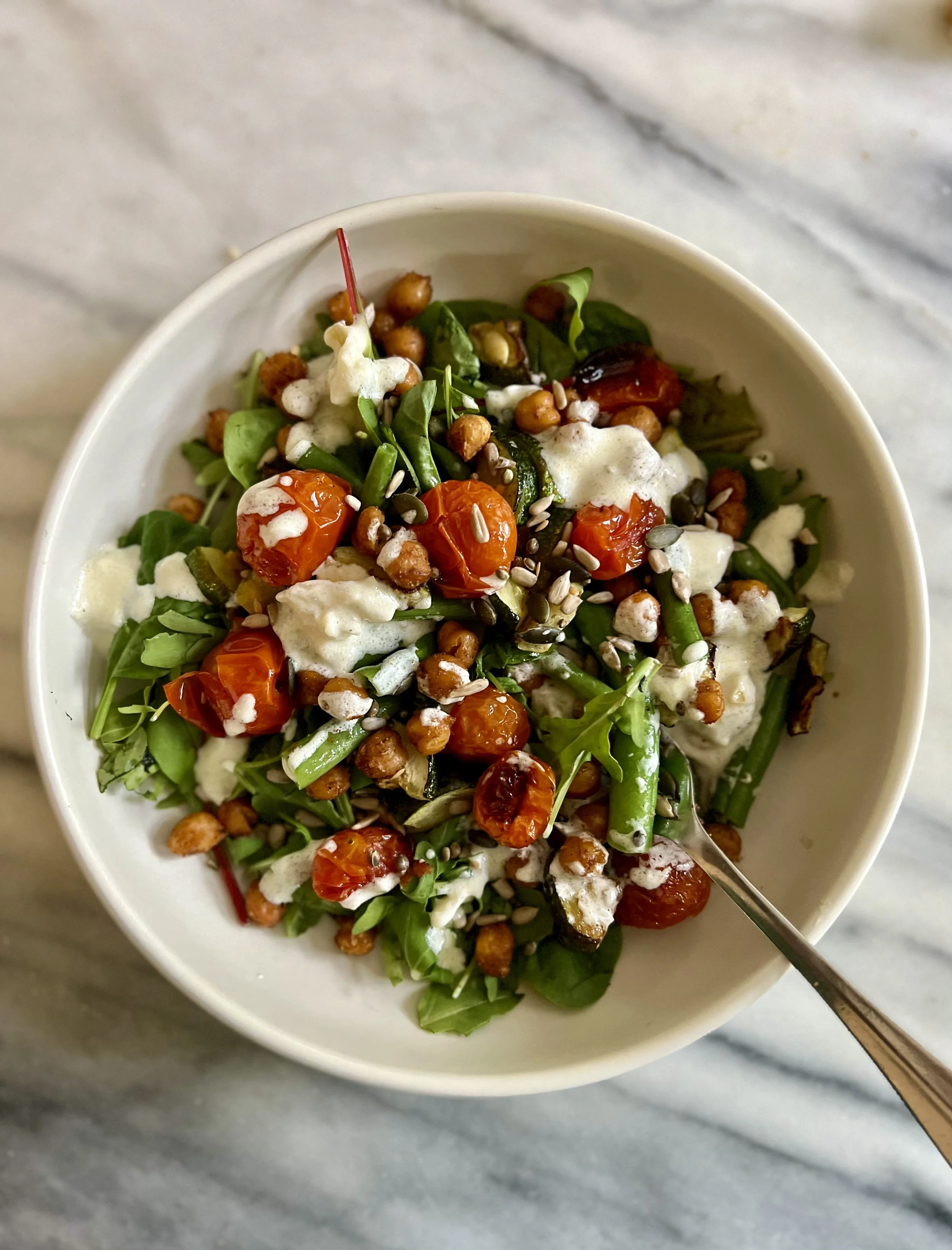 A bowl of mixed greens salad with cherry tomatoes, chickpeas, seeds, creamy dressing, and herbs, on a marble surface with a spoon.