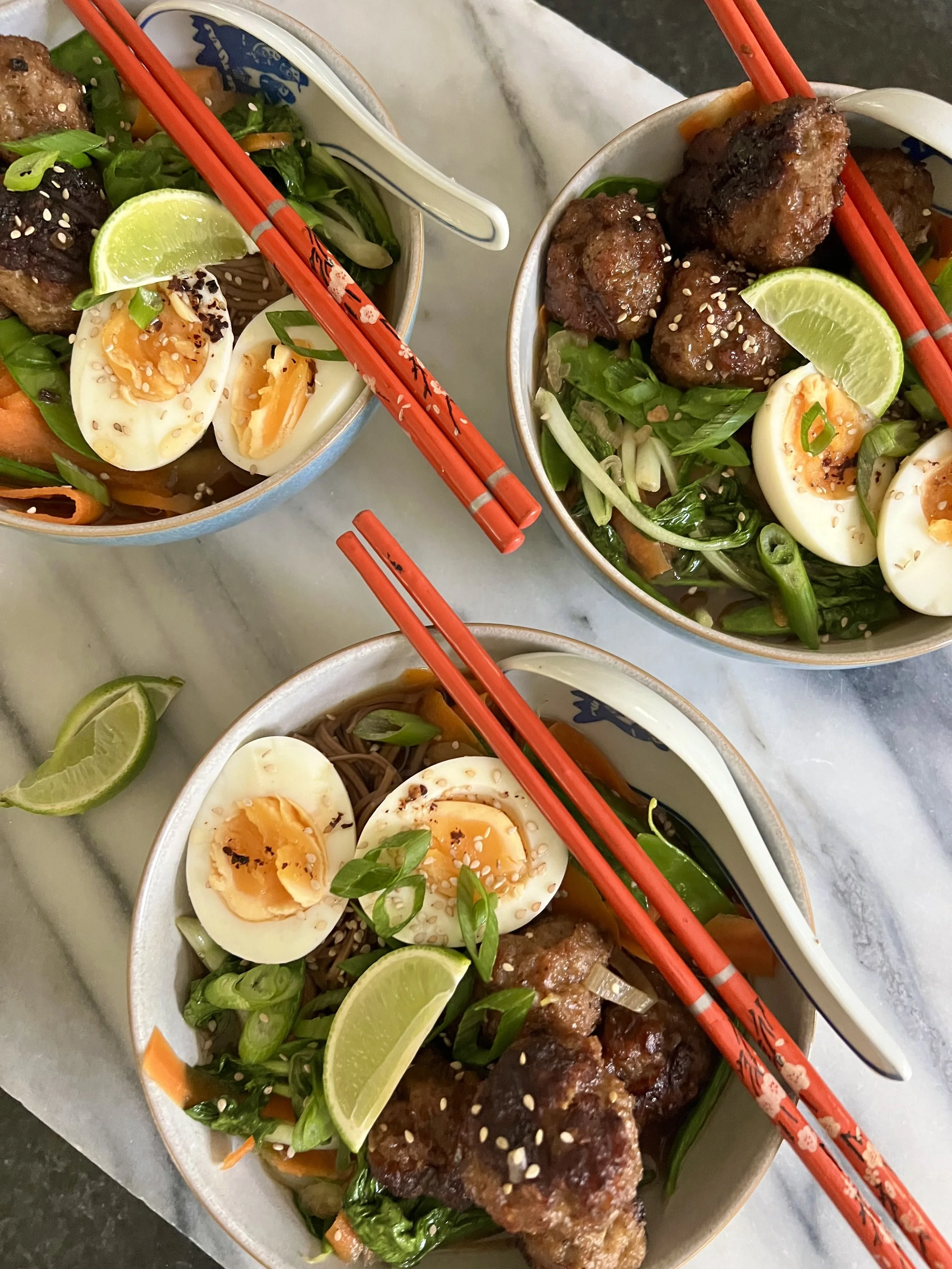 Bowl of Asian-inspired dish with grilled meatballs, sliced soft-boiled eggs, green vegetables, lime wedges, chopped green onions, sesame seeds, and red chopsticks on a light-colored surface.