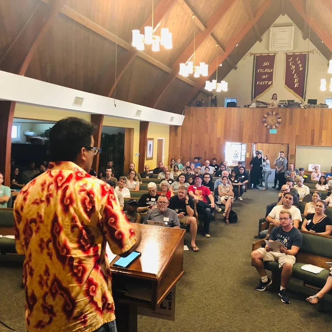 Sam Sukaton in a colorful shirt speaking or presenting in front of a large seated audience in a church or auditorium setting with wooden walls and high vaulted ceiling.