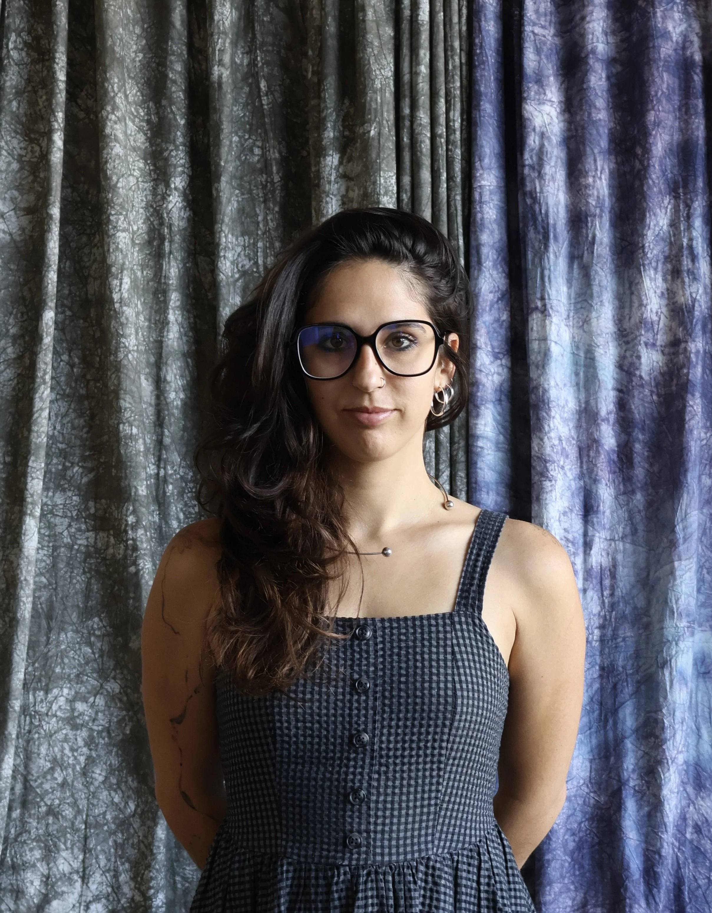 A woman with long dark hair and black-framed glasses wearing a gray button-down dress, standing in front of gray and purple curtains.