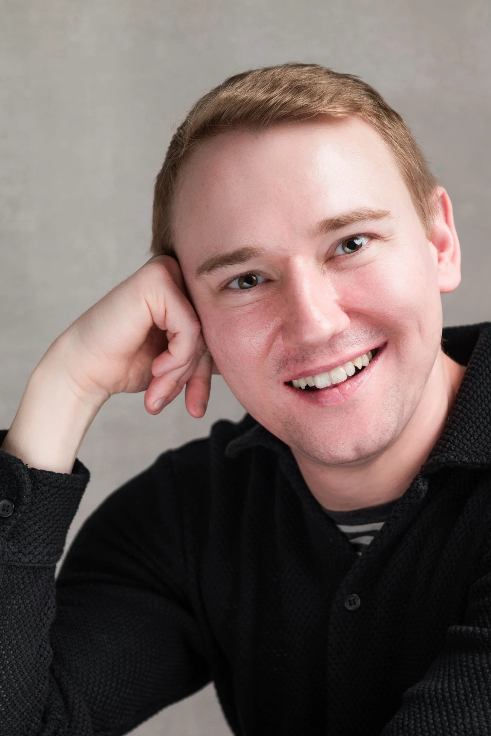 A smiling man with light brown hair resting his cheek on his fist.