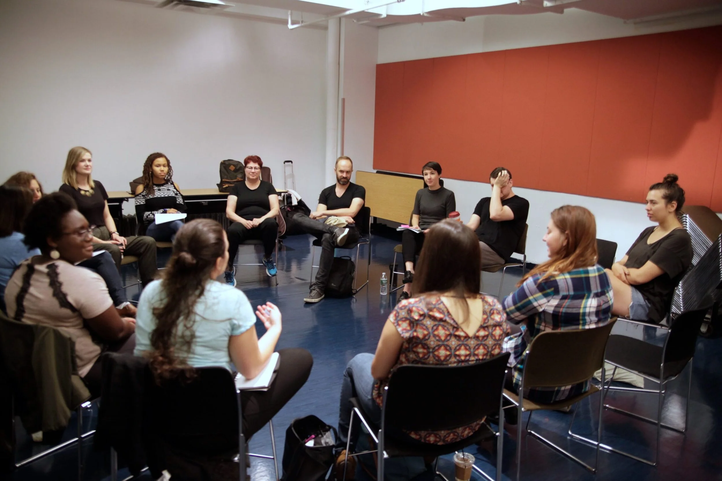 A diverse group of people sitting in a circle in a room with plain walls, engaged in a discussion or workshop.