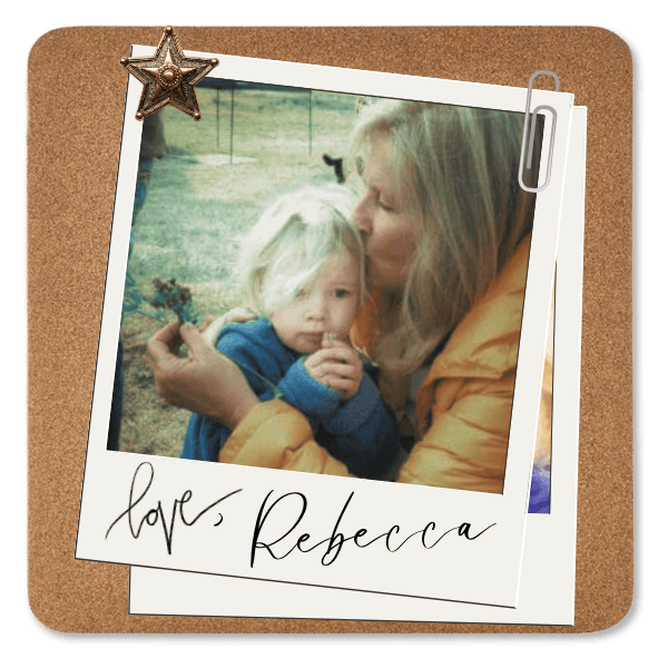 A woman with long, blonde hair wearing a yellow jacket holding a young child with blonde hair wearing a blue jacket outside in a park or yard.