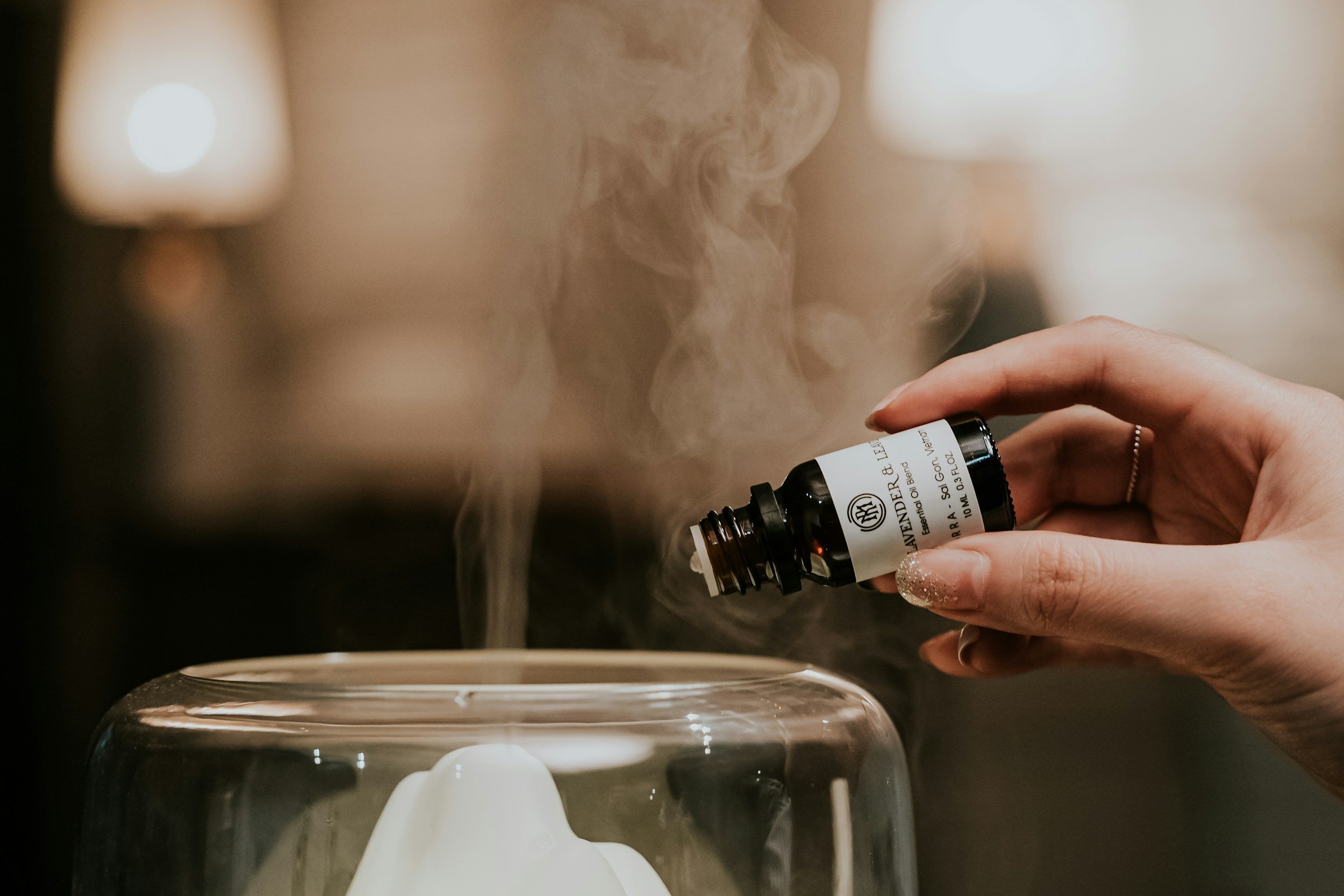 A hand holding a small bottle of essential oil near a diffuser that is releasing steam or fragrance.