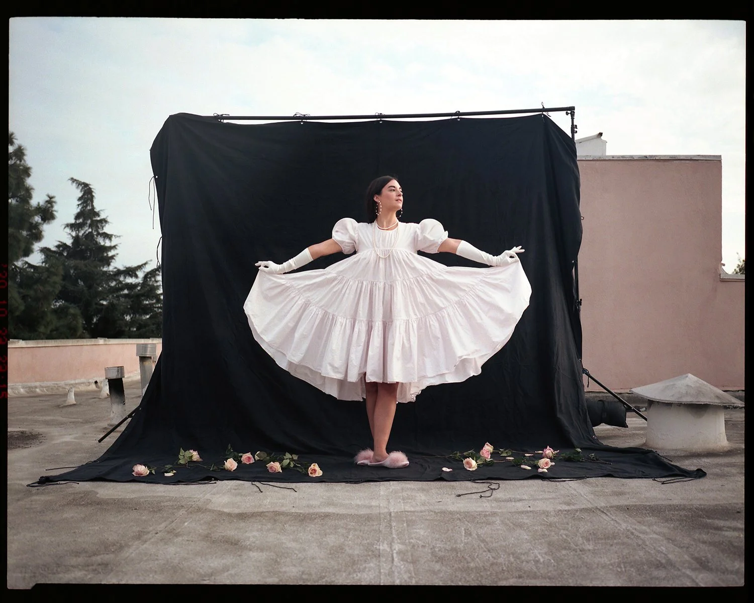 A woman in a vintage white dress with puffed sleeves and gloves poses on a rooftop, lifting her skirt in a ballet stance, surrounded by pink roses and positioned in front of a black backdrop.