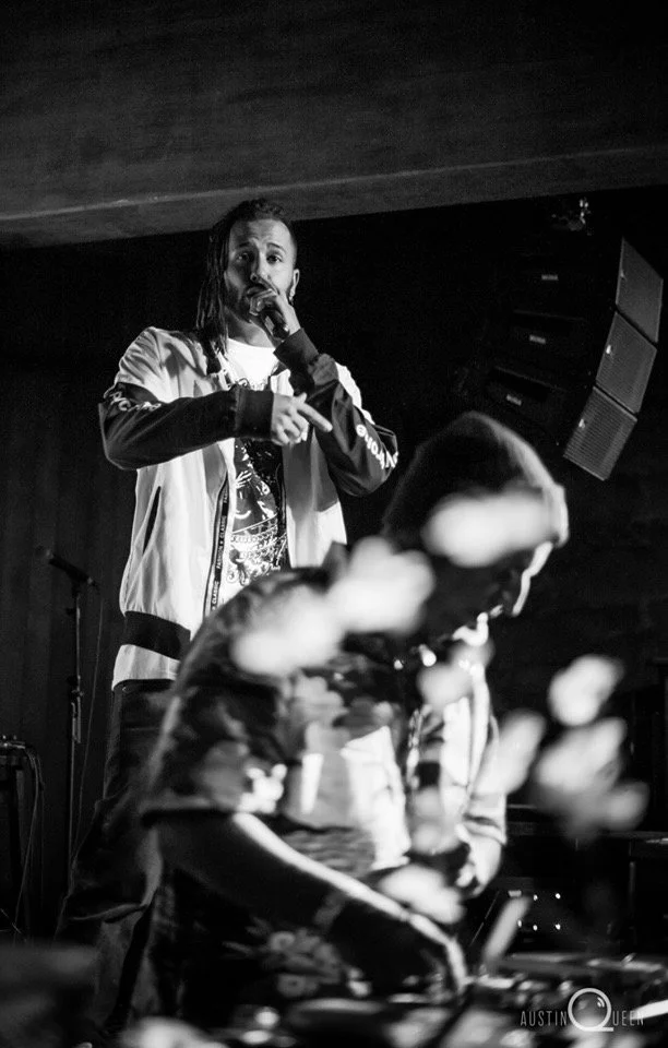 Black and white photo of a male performer on stage holding a microphone, with a DJ in the foreground using equipment, in a dimly lit venue.