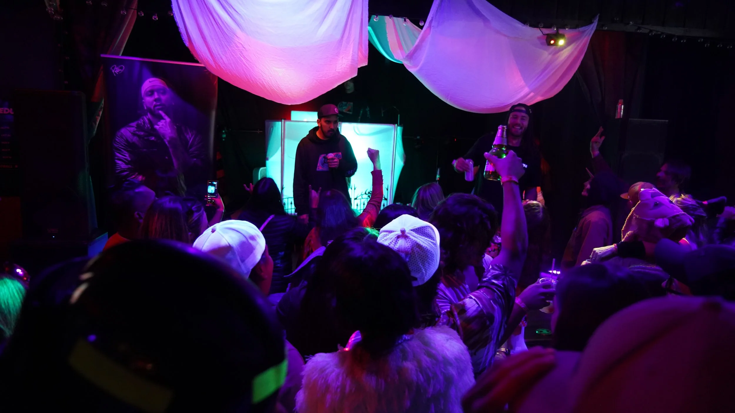 Crowd at a nightclub or concert with colorful lighting and a stage featuring a DJ in the background. Several people are dancing, raising their hands, and holding drinks.