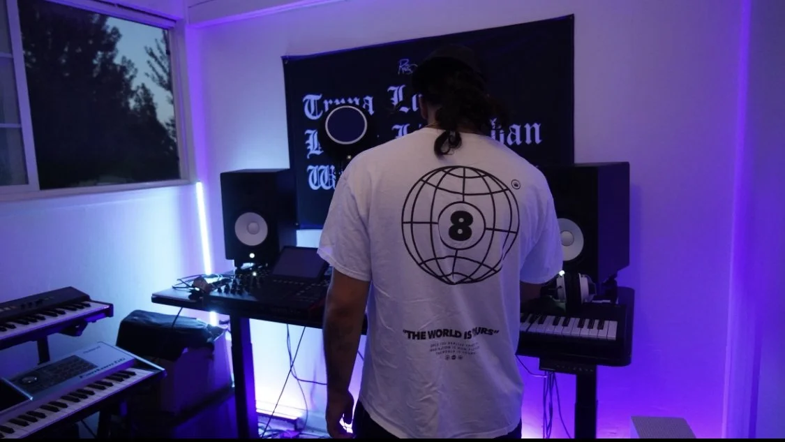 Back view of a person in a white T-shirt with a globe and 8 ball design, in a music studio with electronic keyboards, studio monitors, and a computer, ambient purple lighting.