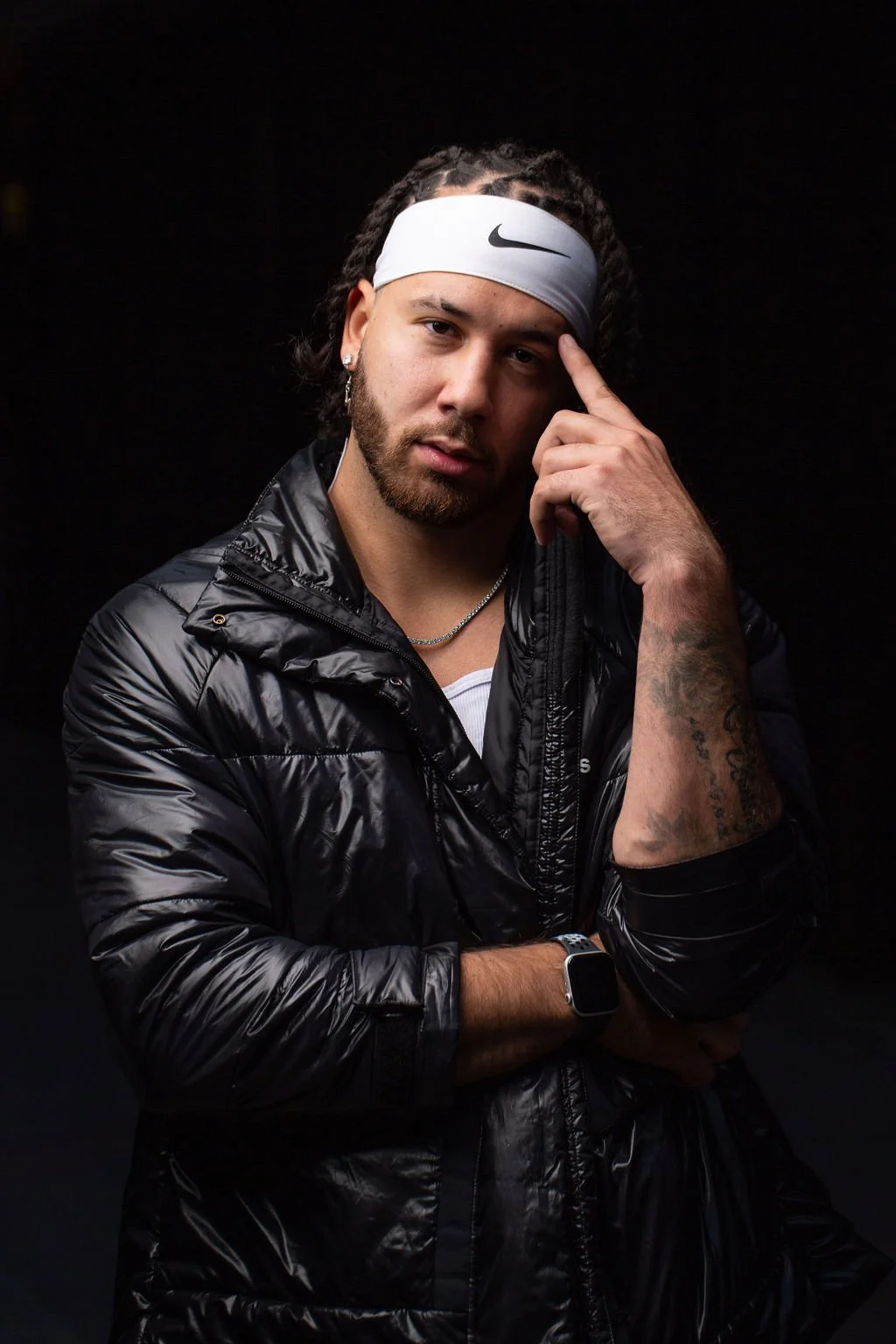 A young man with dark hair and a beard, wearing a white Nike headband, black puffy jacket, and jewelry, poses against a dark background, touching his forehead with his right hand.