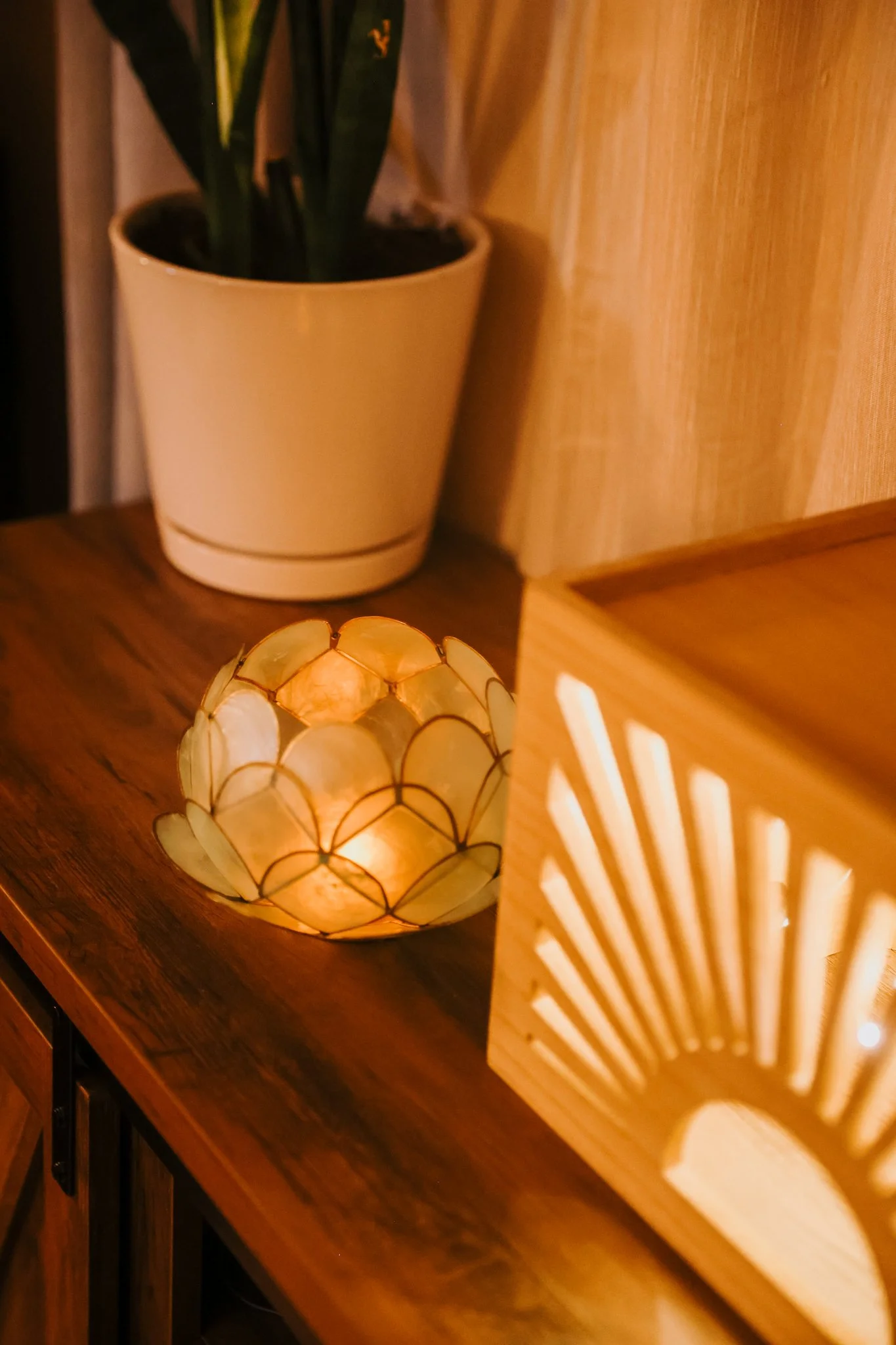 A lit decorative candle holder with petal-shaped pieces on a wooden surface, a potted plant in a beige pot, and a wooden lantern casting a patterned shadow.