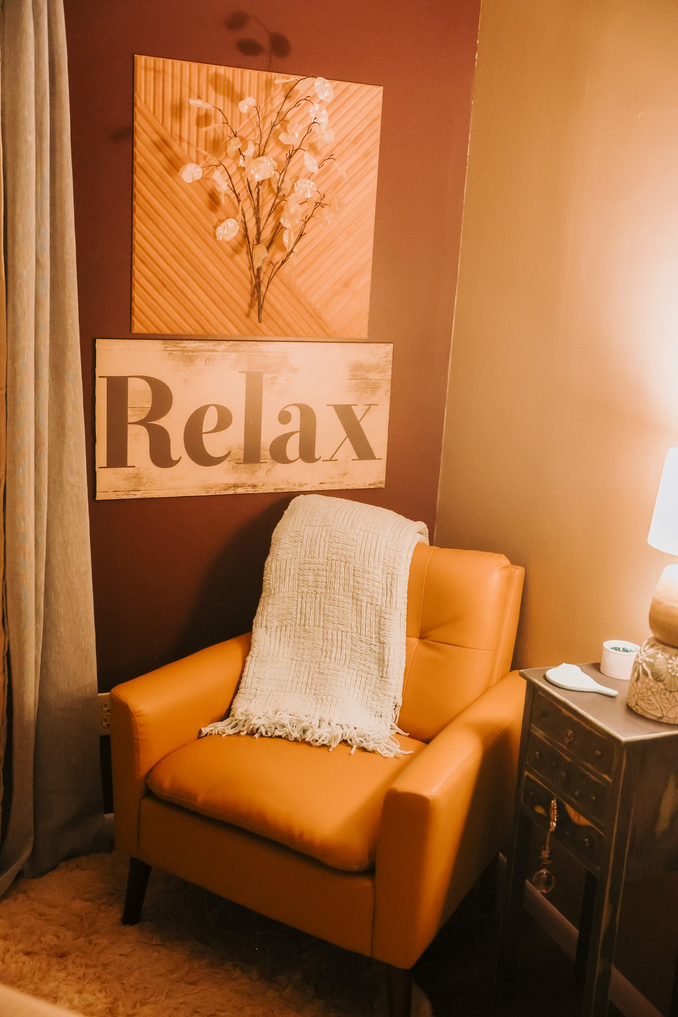 A cozy corner featuring an orange armchair with a beige textured throw draped over it, next to a small dark wood side table with a lamp, candles, and decorative items. Behind the chair, there's a wall with two wooden wall hangings, one with a floral design and the other with the word "Relax" in large letters.
