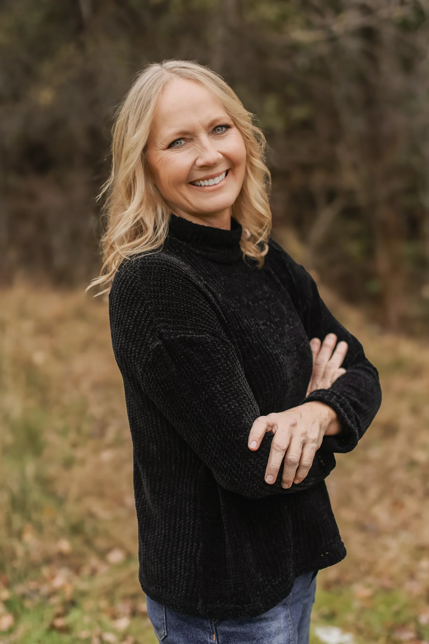 A smiling woman with blonde wavy hair wearing a black sweater stands outdoors with her arms crossed, in front of a blurred natural background.