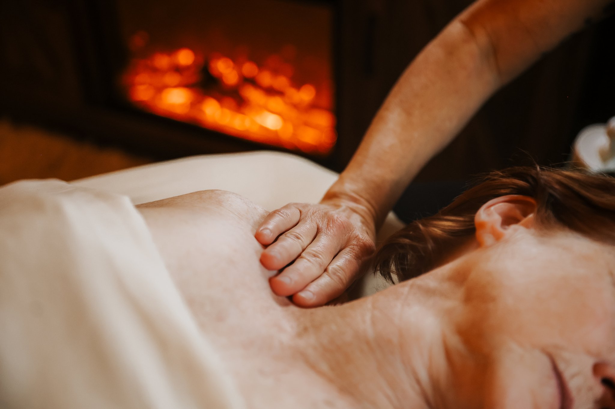 A person with tattoos receiving a massage with their head turned to the side, revealing a tattoo behind their ear and short hair, lying on a massage table.
