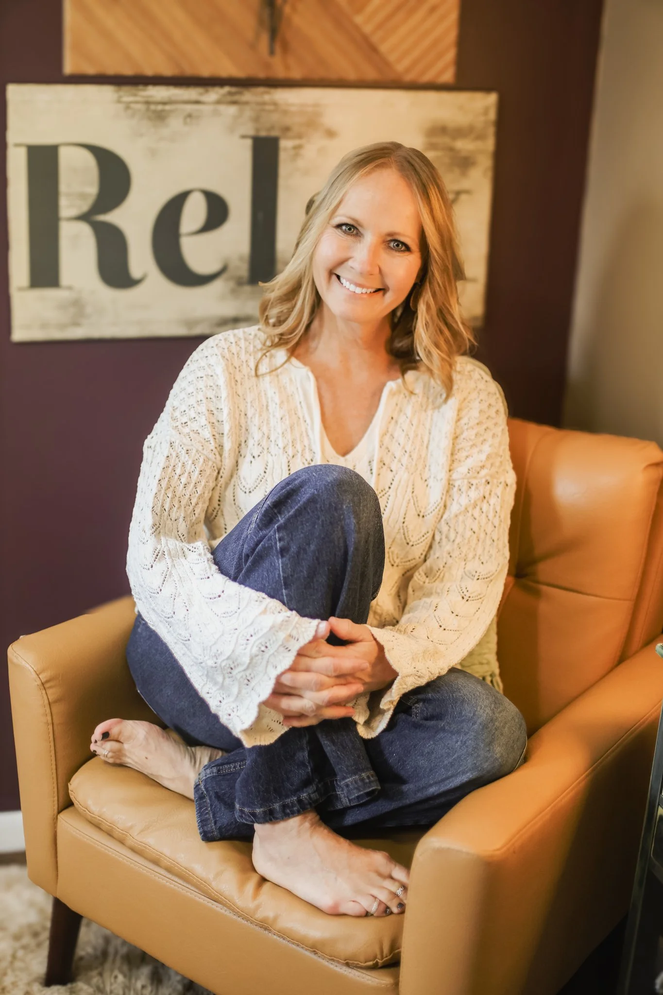 A woman with blonde hair, wearing a light-colored lace sweater and blue jeans, sitting cross-legged on a tan armchair inside a room. She is smiling and looking at the camera. Behind her is a wall with a sign that reads 'Relax'.