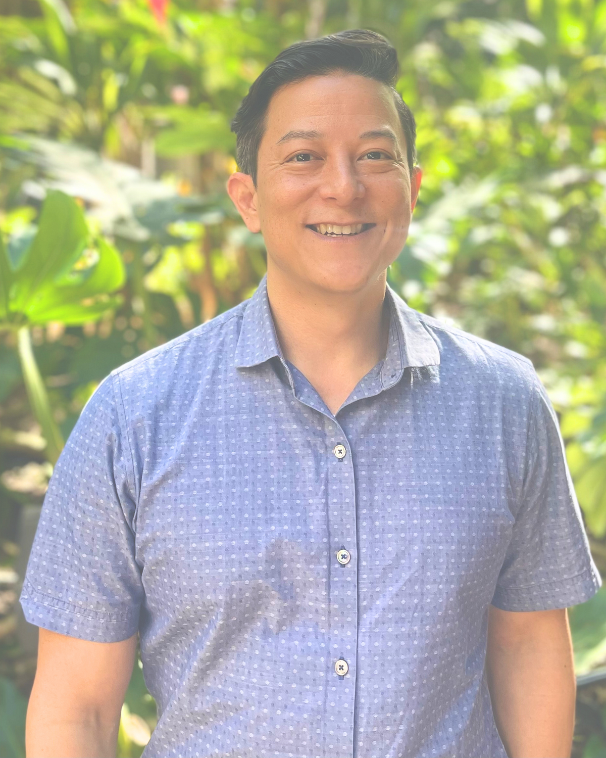 A smiling man with dark hair wearing a light blue, short-sleeved, button-up shirt standing outdoors with green foliage background.