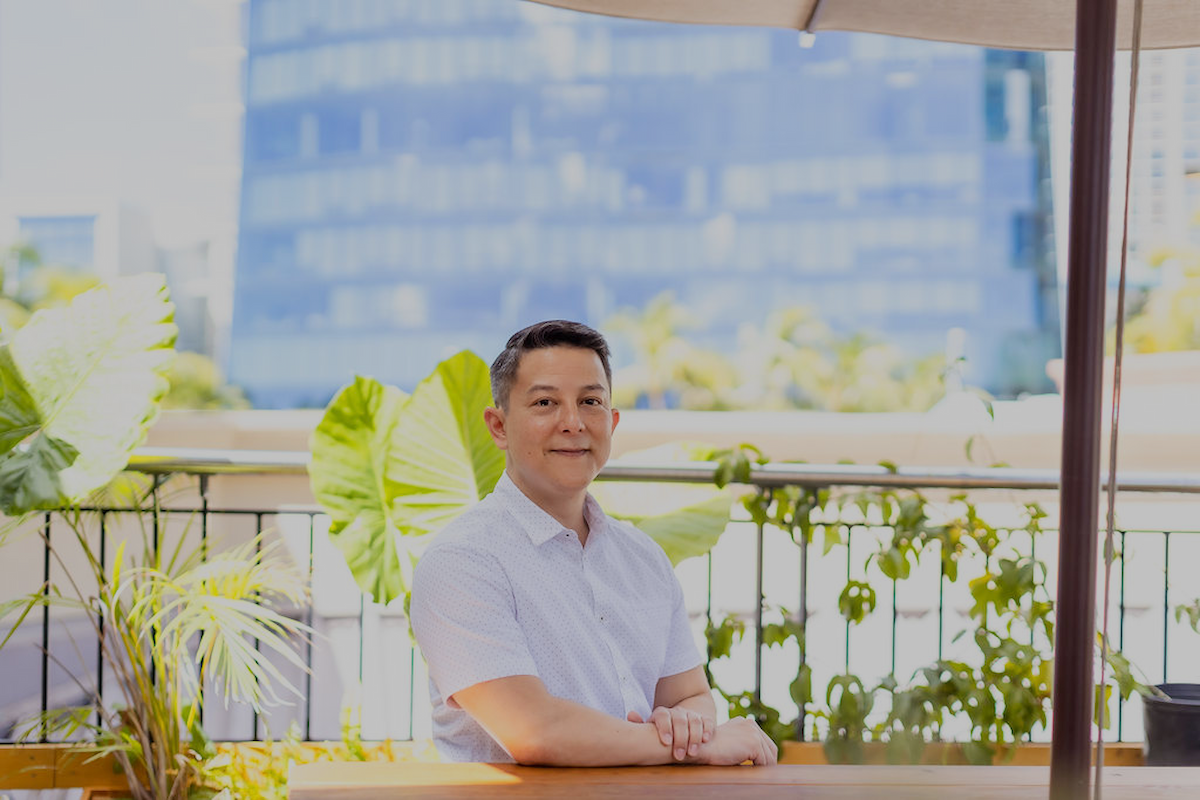A man sitting at a table outdoors on a balcony or patio with green plants in the background, and tall buildings in the distance, during daytime.