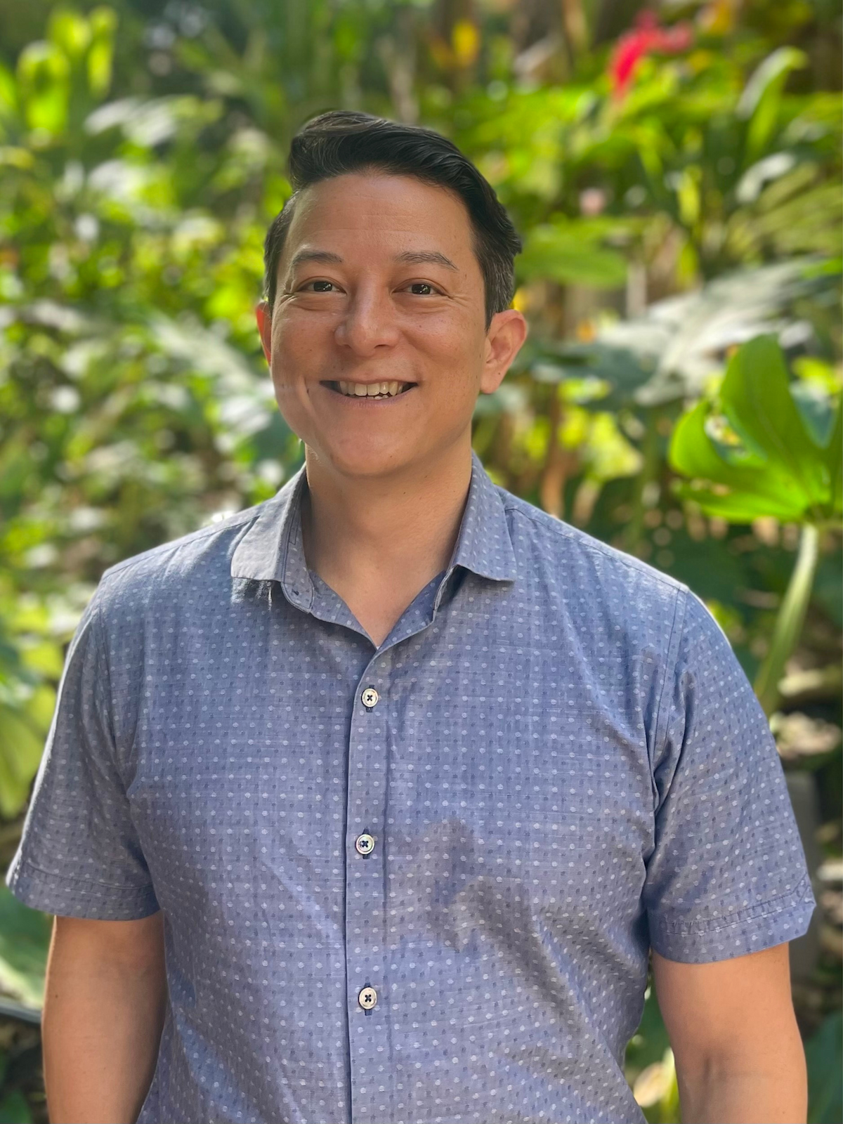A smiling man in a short-sleeved, button-up, blue patterned shirt standing outdoors with lush green foliage in the background.
