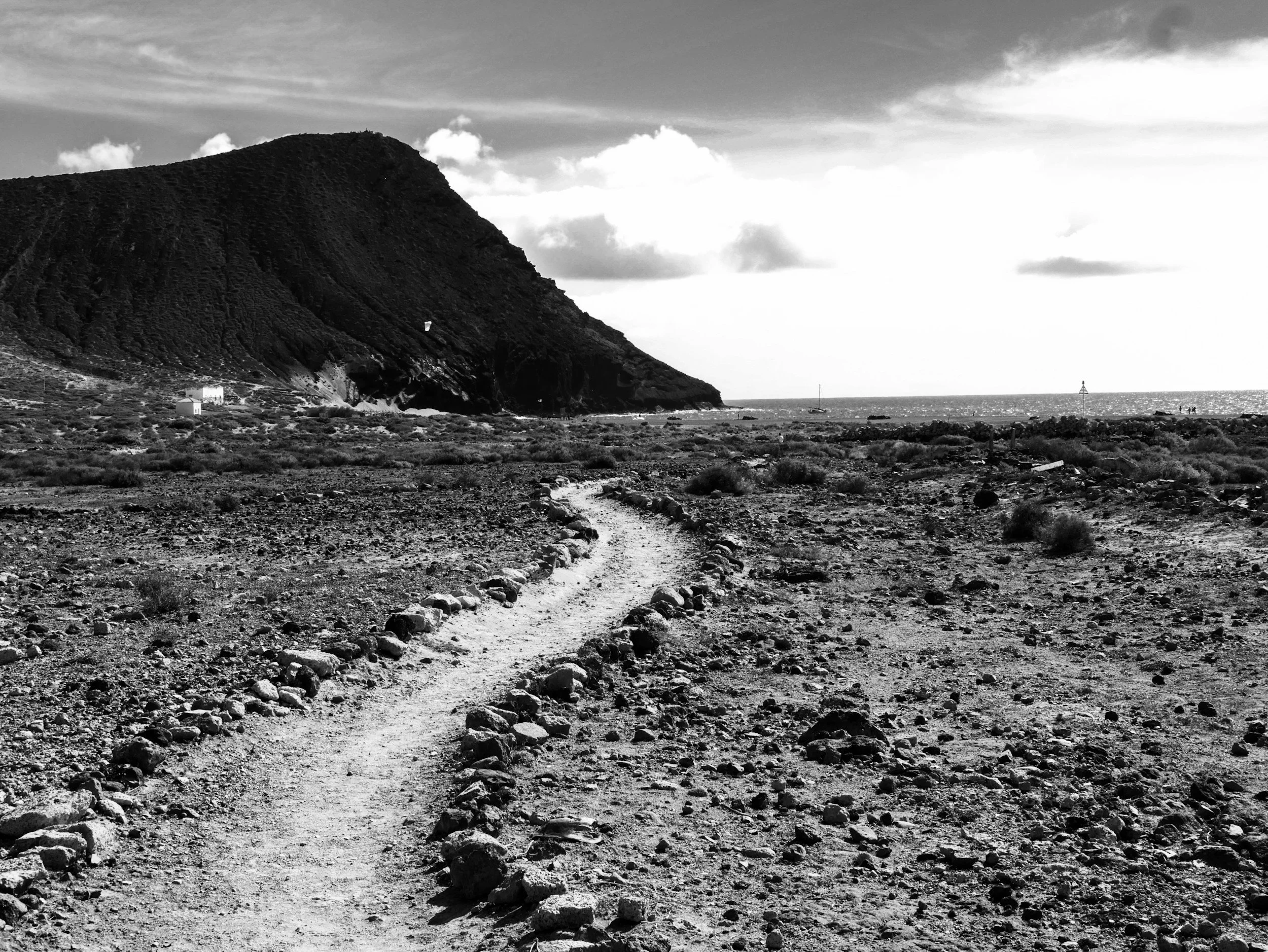 A rugged dirt path winding through a barren, rocky landscape leading towards a large hill or small mountain near the ocean, with scattered shrubs and a cloudy sky overhead.