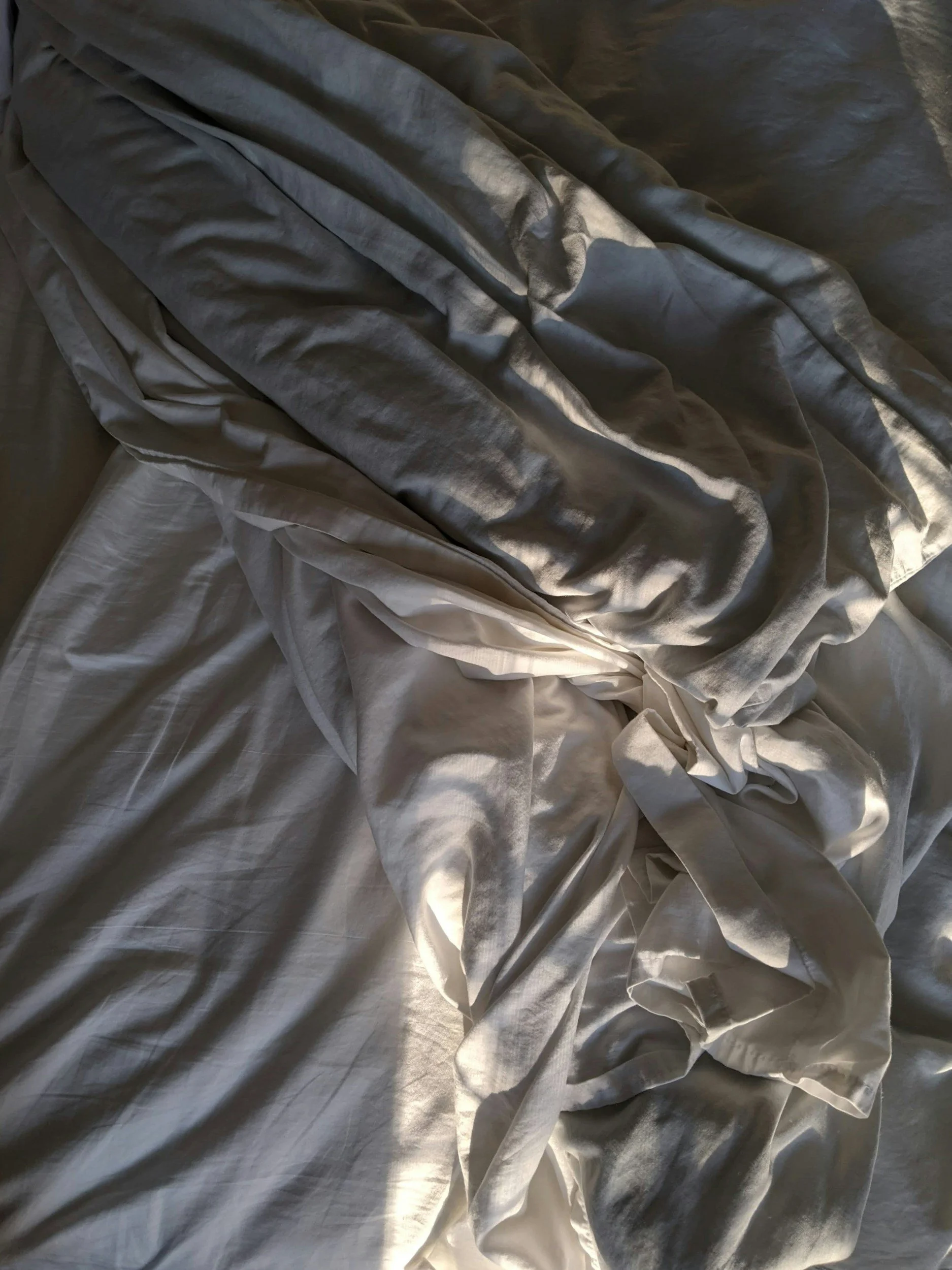 Crumpled bed sheets in shades of silver and white with sunlight creating shadows.