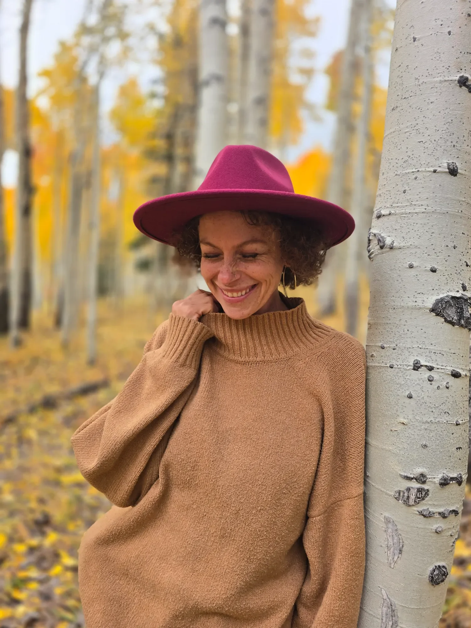 A woman with curly hair, wearing a wide-brimmed pink hat and a tan sweater, smiles with her eyes closed while leaning against a white aspen tree in a forest with yellow autumn leaves.