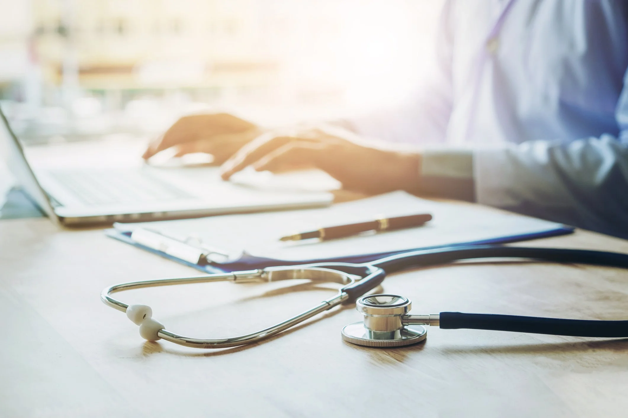 A stethoscope, a pen, a clipboard, and a laptop on a wooden desk with a physician, doctor, nurse practitioner, wellness professional in a white lab coat working on the laptop in the background.