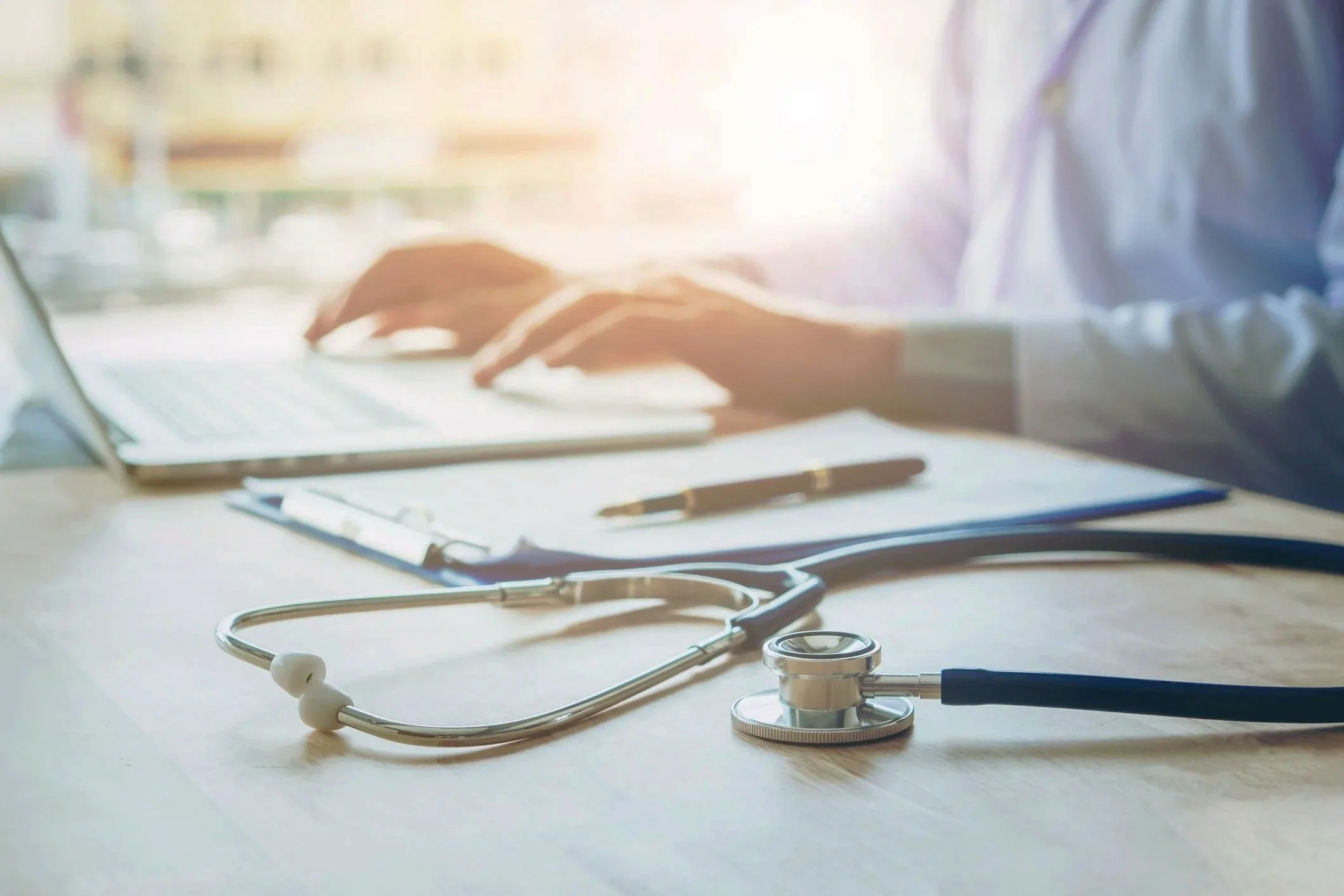 A stethoscope on a wooden table with a clipboard, pen, and physician or nurse practitioner working on a laptop in the background.