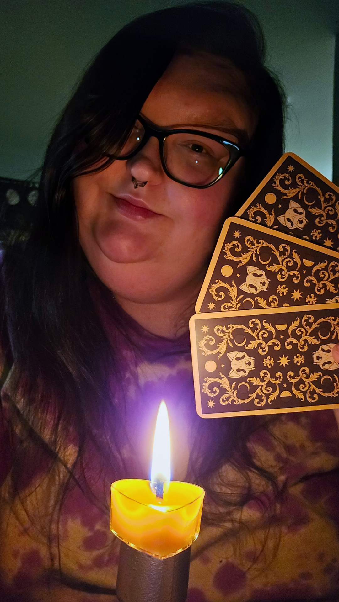 Person with long dark hair, glasses, and a septum piercing holding three decorated tarot cards and a lit candle in front of them in a dark room.