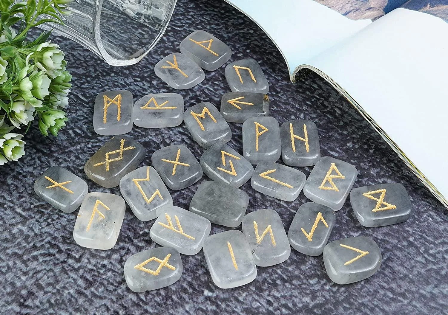 A collection of rune stones with gold inscriptions scattered on a dark textured surface, accompanied by a partially open book, a glass of water, and a small bouquet of white flowers.