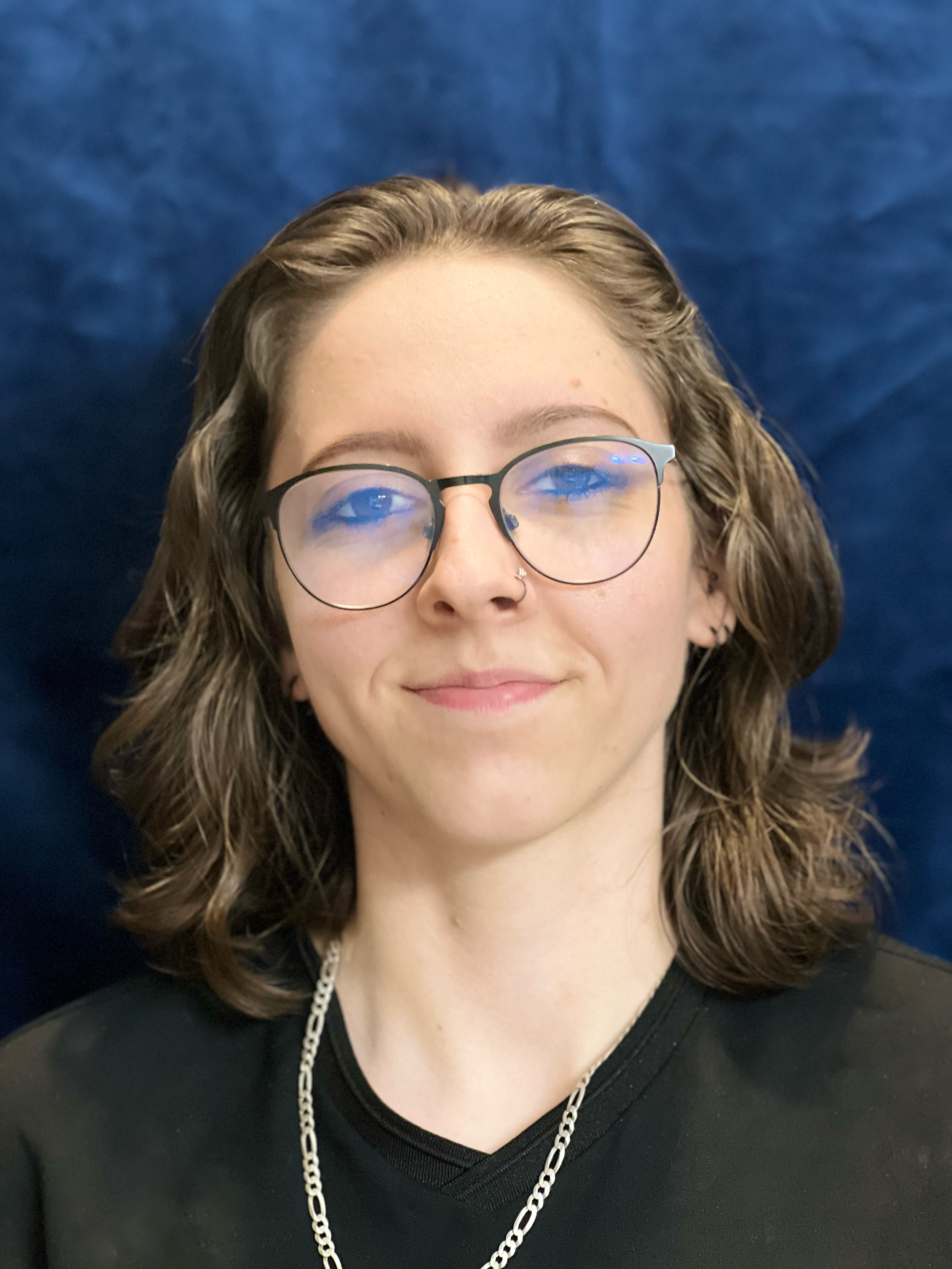 A woman with shoulder-length wavy brown hair, glasses with round black frames, a nose ring, and a silver chain necklace, smiling softly and wearing a black shirt against a blue background.