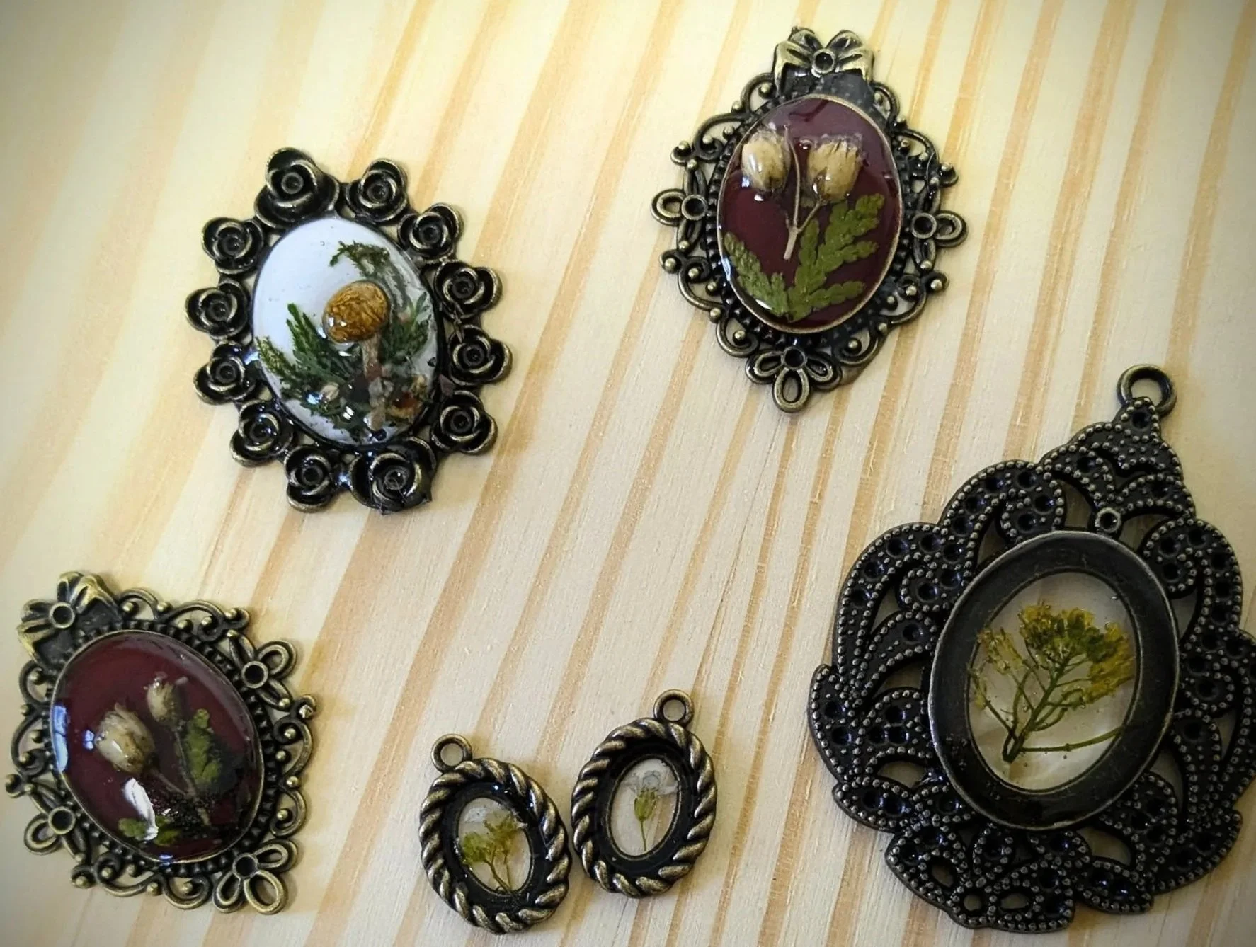 Decorative jewelry pendants with pressed flowers and herbs in vintage-style metal frames, displayed on a wooden surface.