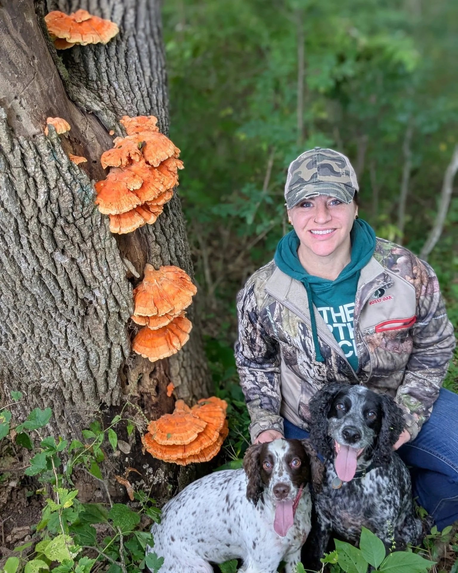 A woman dressed in camouflage and outdoor clothing kneeling beside two dogs, one black and white and the other brown and white, in a wooded area. There are orange mushrooms growing on a tree trunk next to them.